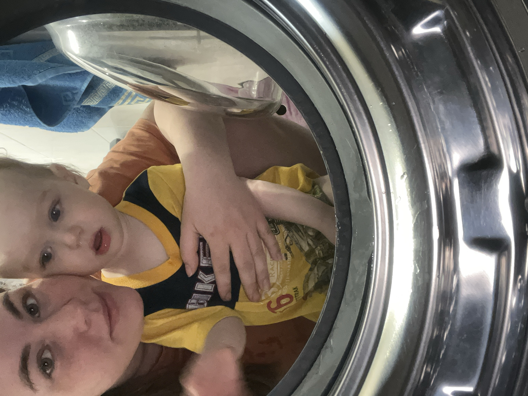 A photo taken from inside a washing machine depicts a woman and a young child. The woman, positioned at the bottom of the frame, is looking towards the camera. A child is nestled against her, wearing a yellow t-shirt with a black collar and an emblem on the front. The setting appears to be domestic, with a blue towel visible in the background. The location is Chaikovsky, Russia.