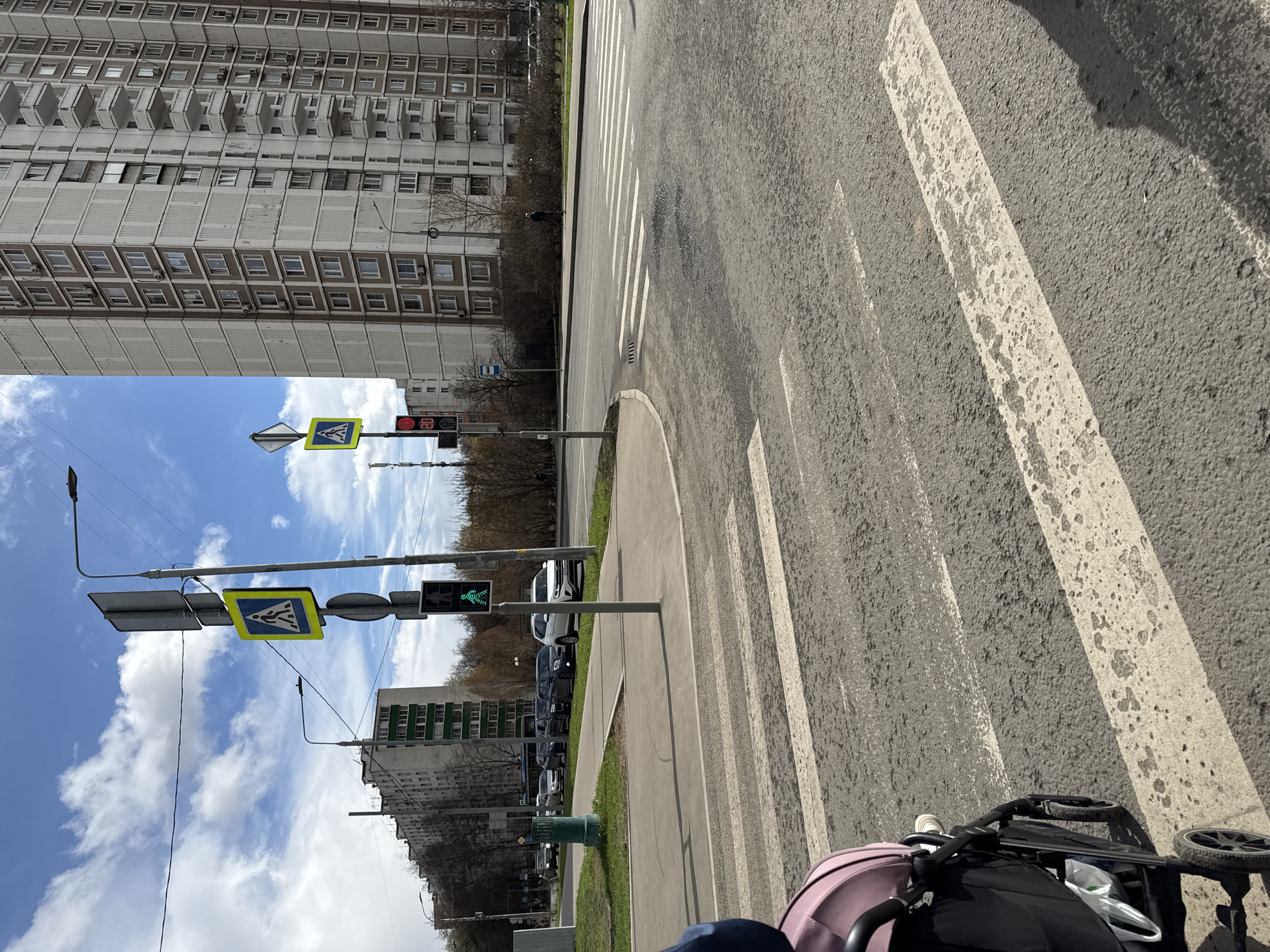 A street scene in Moscow, Russia, shows a pedestrian crossing with white stripes on a gray asphalt road. A tall, Soviet-era apartment building with rows of balconies looms in the background. Traffic signals display a green light for pedestrians. A stroller is visible in the foreground, partially obscuring the view. Cars are parked along the side of the road. The sky is partly cloudy with blue patches.