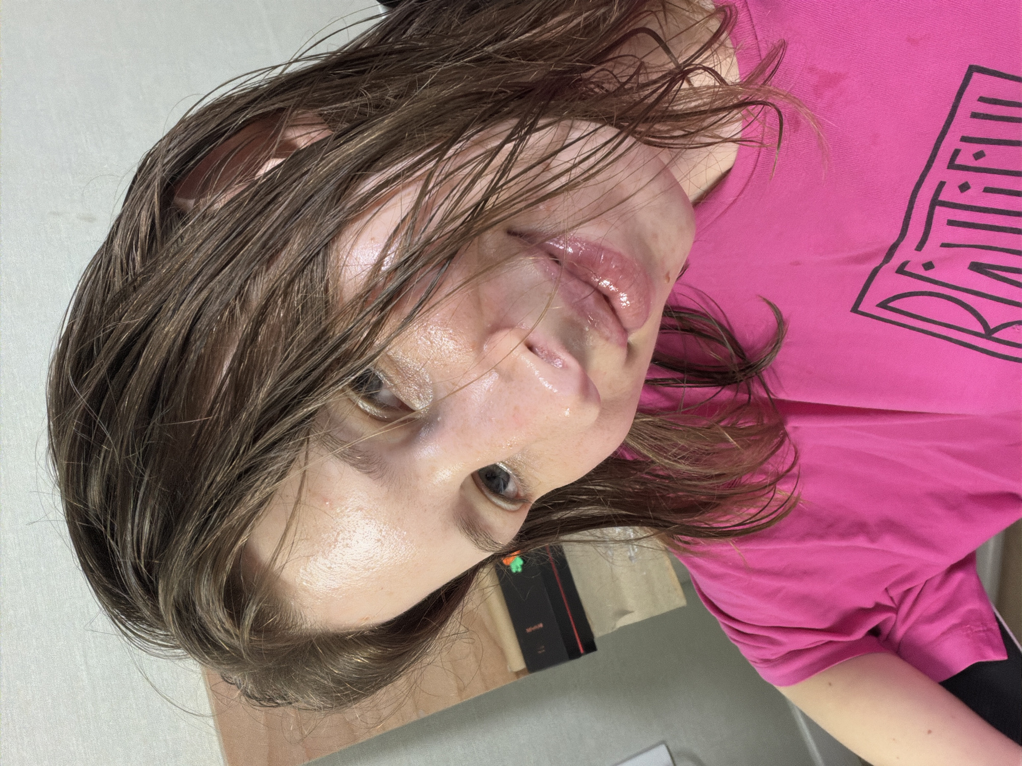A young woman with wet, brown hair and light skin is captured in a close-up selfie taken indoors. She is wearing a bright pink t-shirt with a graphic design on it. Her eyes are open, and her head is tilted, with strands of hair falling across her face. The background appears to be a wall with a wooden surface behind her to the left. There are no other individuals or clear signs of the specific location within Dongducheon-si, South Korea. The overall impression is of an informal, personal photograph, possibly taken after an activity that caused her hair and face to be wet.