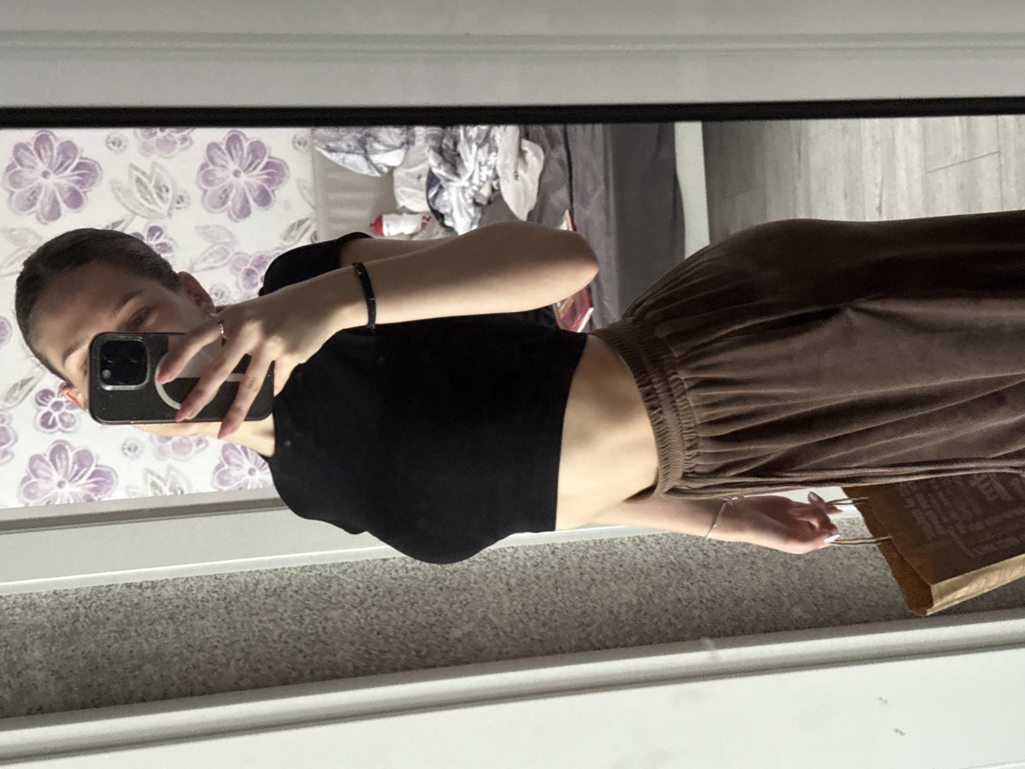 A young woman is captured in a mirror selfie, standing upright and viewed from the waist up. She has dark hair styled back, and her face is largely obscured by the dark-colored smartphone she holds in her right hand, pointed towards the mirror.
She is wearing a fitted black crop top that reveals a sliver of her midriff, paired with brown, high-waisted, wide-leg pants that appear to be made of a soft, possibly velour or ribbed fabric, featuring an elasticated waistband. Accessories include a thin silver ring on her right hand, a black beaded bracelet on her left wrist, and a delicate silver bracelet on her right wrist. Her nails have a light polish. In her right hand, near her leg, she also holds a brown paper bag with handles, on which some dark, illegible text is visible.
The background, a reflection in the mirror, depicts a domestic setting. To the left, there's a light-colored wall adorned with a floral wallpaper featuring purple and green flower patterns. To the right, a pile of clothes and other items are visible, possibly on a bed or chair, against a backdrop of grey curtains or fabric. The floor in the foreground of the reflection shows a light grey carpet, while behind her legs, a light-toned wooden or laminate floor is reflected. The mirror itself is framed by white trim.
The activity is clearly a mirror selfie, taken casually indoors. There are no specific cues for time of day or weather, as it's an indoor shot. The overall scene suggests a personal space, likely a bedroom or dressing area, in Sofia, Bulgaria.