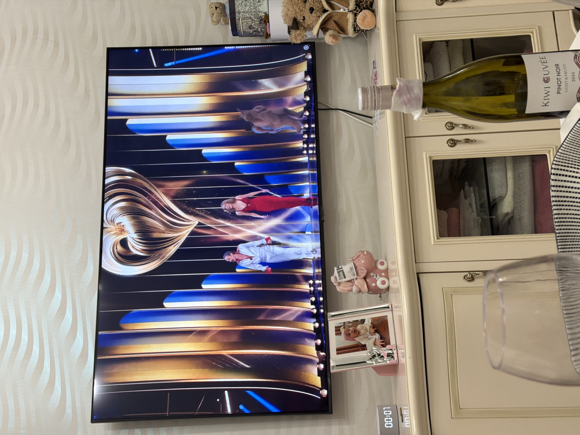 A detailed, matter-of-fact description of the image:
The image appears to be taken in an indoor residential setting in Khimki, Russia. A flat-screen television is mounted on a wall with patterned wallpaper, displaying a scene of individuals in formal attire; one woman is wearing a red dress and a man is wearing a white suit, against a backdrop of swirling gold and blue lines. Above the television are several small decorative items including teddy bears and a small decorative box. To the right of the television is a light-colored cabinet with glass-paneled doors, visible with towels and textiles inside. A bottle of "KIWI CUVEE" Pinot Noir wine and a wine glass are on top of the cabinet. Also visible are a pink piggy bank reading a newspaper, a small framed photo (possibly of a child), and a digital alarm clock. The presence of these items indicates that the location is in a private dwelling in Khimki, Russia.