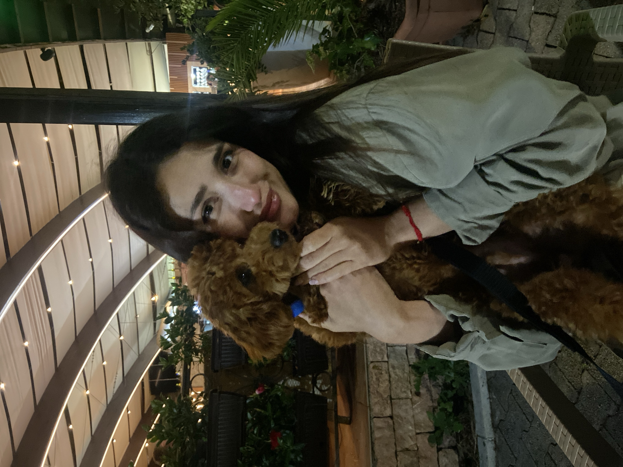 A woman with long dark hair is holding a brown poodle. The poodle has a blue collar. The woman is wearing a light green shirt and a red string bracelet on her left wrist. They are outside at night. In the background is a building with a curved, illuminated roof and potted plants. The location is Bar, Montenegro.