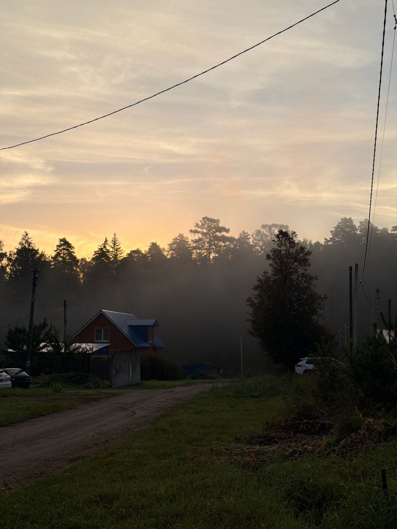 This is a photograph taken at dawn in Chaikovsky, Russia. The sky is filled with soft, hazy clouds tinged with the orange glow of the rising sun. A thin power line stretches diagonally across the upper portion of the frame. In the mid-ground, a two-story house with a red facade and a blue roof is visible. It is partially obscured by mist and trees. A dirt road leads towards the house from the lower left of the image. The surroundings are a mix of trees, some of which are silhouetted against the sky, and overgrown grass. Two cars are parked near the house. Electrical poles and wires are present throughout the scene. The overall atmosphere is serene and slightly melancholic, characteristic of early morning fog in a rural or semi-rural setting.
