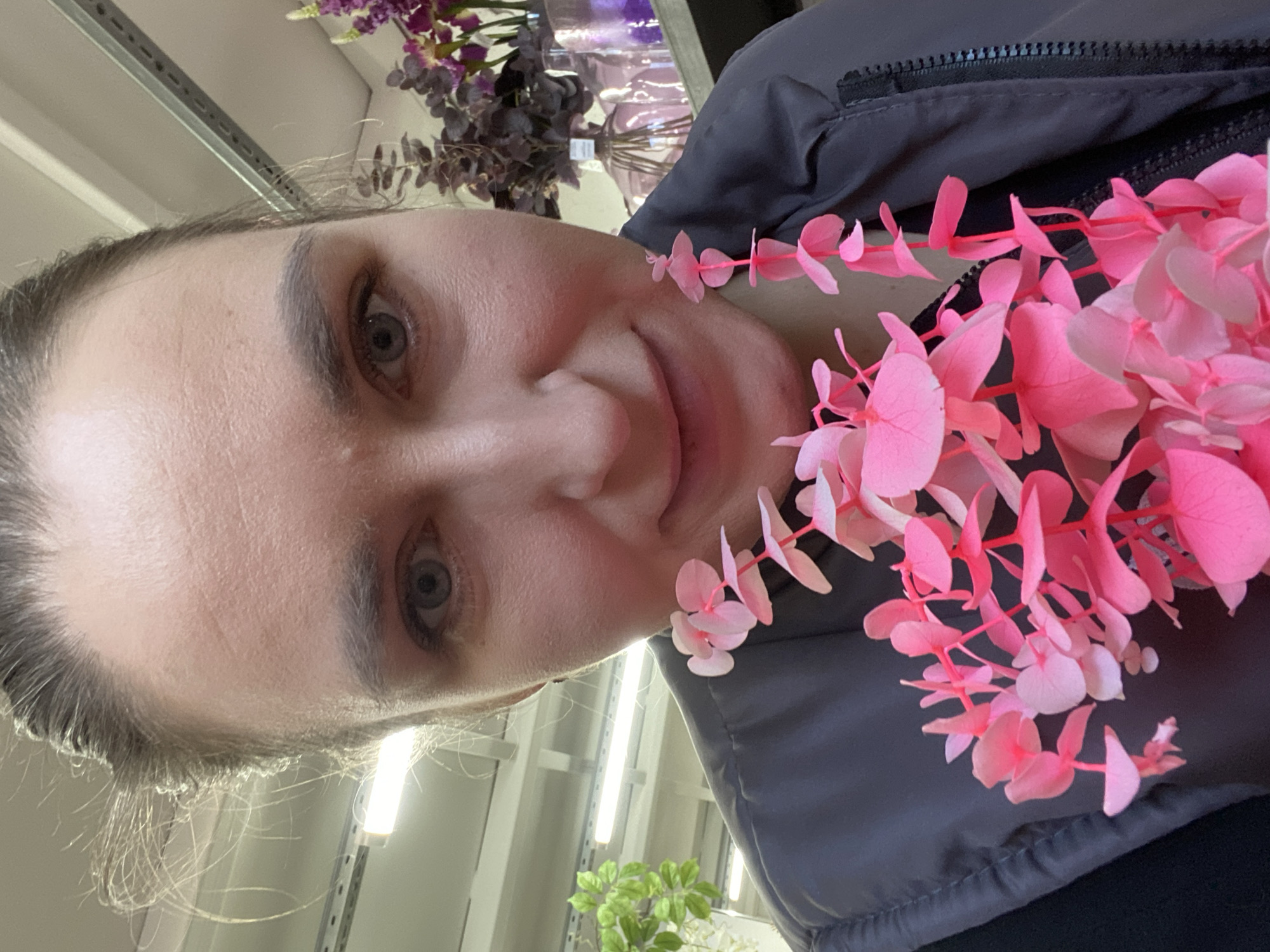 A selfie taken at an upward angle shows a woman with light skin, gray eyes, and dark hair. Her face is the primary subject, occupying the left side of the frame. She is wearing a dark gray vest. Around her neck is a strand of artificial pink eucalyptus leaves. In the background, there are rows of fluorescent lights, suggesting an indoor commercial space, likely a store or warehouse. Above the woman, artificial purple and green plants are visible on a shelf. The location is consistent with a retail establishment in a city like Yekaterinburg, Russia.