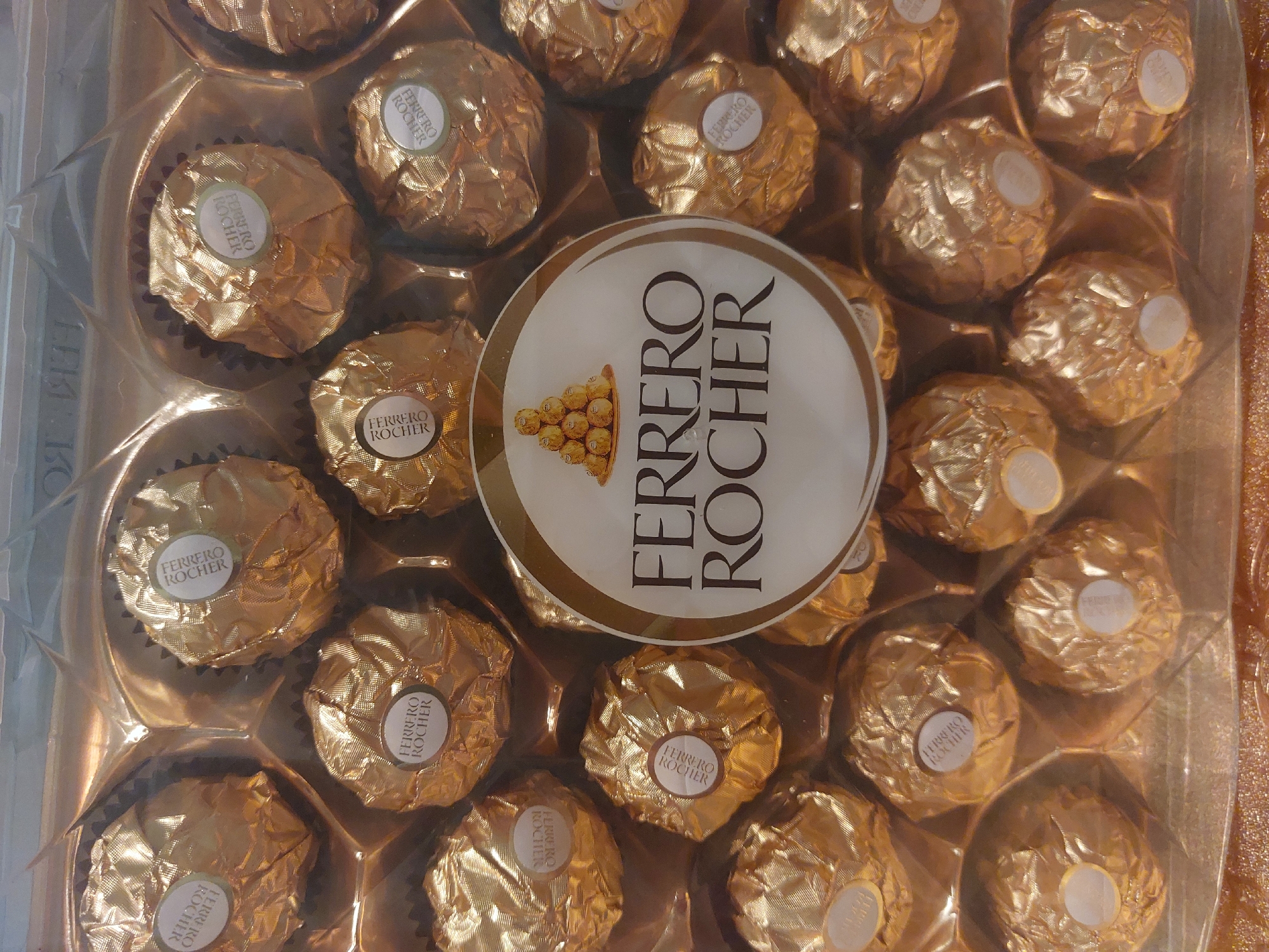 The image shows a plastic container filled with Ferrero Rocher chocolates. The chocolates are individually wrapped in gold foil and have a round sticker with the Ferrero Rocher logo on them. A circular label with "Ferrero Rocher" printed on it covers some of the chocolates. The plastic container is clear and shows the chocolates inside. The background is blurred, but it appears to be a reflective surface, possibly a table. There is no indication of a location within Tsarevo, Bulgaria, or any people, subjects, actions, or interactions present in the image.