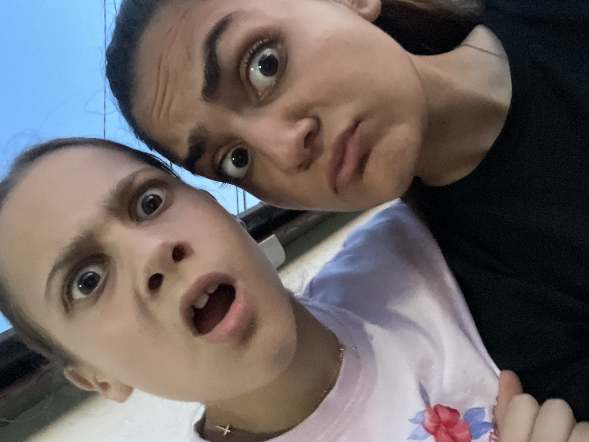 Two young individuals are captured in a close-up, frontal shot, displaying exaggerated facial expressions outdoors in the city of Debelt, Bulgaria. The person on the left, partially cropped, presents a wide-eyed, open-mouthed expression with visible upper teeth. This individual wears a light-colored t-shirt with a red floral design, a thin gold-colored chain necklace, and a small gold-colored cross earring in the left ear. The person on the right, more centrally positioned, also exhibits a wide-eyed expression but with a closed mouth, lips pursed downwards in a pout. This individual has dark hair pulled back and wears a dark-colored t-shirt with a thin gold-colored chain necklace. Both individuals are looking directly at the camera. The background is out of focus, revealing a clear blue sky in the upper right and a light-colored architectural surface in the upper left, with a thin dark line discernible against the sky. The lighting suggests ambient outdoor conditions.