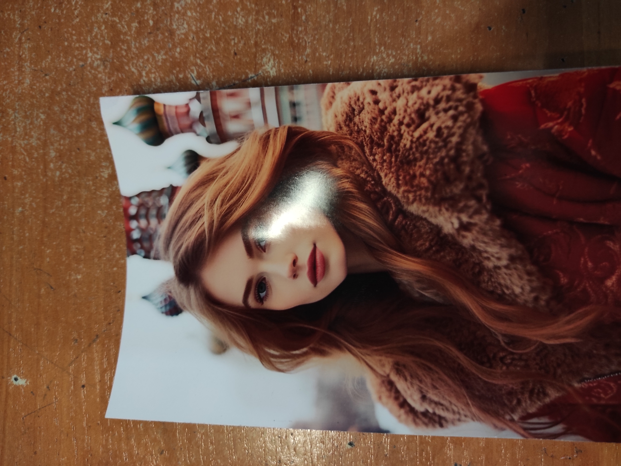 A portrait shows a young woman with long, wavy auburn hair. She is wearing a red coat with a fur collar. The background features the colorful domes of a building that resembles Saint Basil's Cathedral in Moscow, Russia. The image is a photograph printed on paper, placed on a wooden surface. There is a bright light glare across the woman's face. The location is not Melitopol, Ukraine.