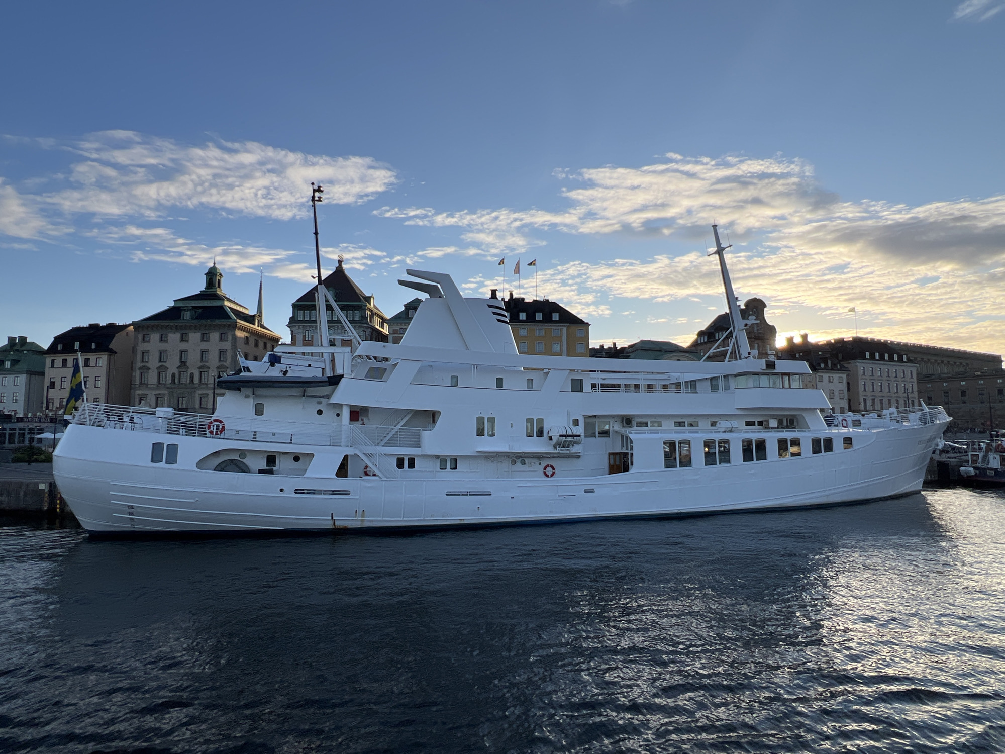 The image shows a large, white cruise ship docked in a harbor. The ship is facing left and its hull is partially submerged in dark blue water. The background features a cityscape with several historic buildings of varying heights and architectural styles, with some featuring green roofs. A flag, appearing to be the Swedish flag, is visible on the left side of the ship. The sky above is blue with scattered white clouds, and the sun appears to be setting on the right side of the image, casting a warm light on the scene. The overall impression is of a calm, late afternoon or early evening in an urban harbor setting, likely Stockholm, Sweden, given the presence of the Swedish flag and characteristic architecture.