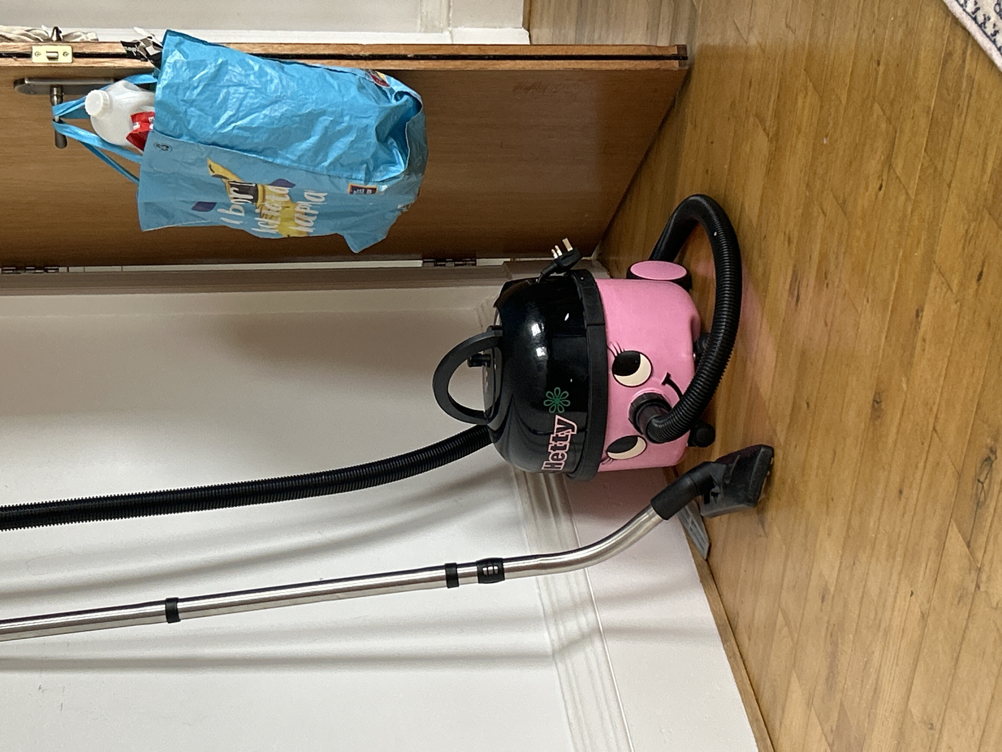 An upright vacuum cleaner with a chrome extension wand, a black flexible hose, and a pink character-like vacuum body with black detailing is positioned on a wooden floor next to a white wall. The vacuum cleaner has a black top with a handle and a black power cord plugged into an unseen outlet. To the right of the vacuum, a wooden door is ajar, revealing a white wall and part of a doorway on the right. A blue reusable shopping bag filled with items, including a white plastic bottle, is hanging from the door handle. The floor is made of polished wooden planks. The scene appears to be indoors, likely in a domestic setting. The image does not provide any information about the City of Edinburgh, United Kingdom.
