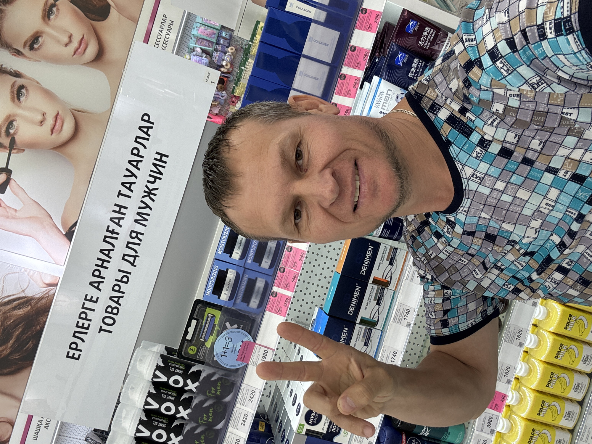 The image shows a man in a store, likely a supermarket or pharmacy, in Oskemen, Kazakhstan. He is taking a selfie with a display of men's grooming products behind him. The man is wearing a checkered shirt and has a silver chain around his neck. His right hand is raised, showing two fingers in a peace sign. The shelves behind him are stocked with various products, including shaving cream, moisturizer, and lotions, some with labels in Russian and possibly Kazakh. There is a large sign at the top of the display that reads "ЕРЛЕРГЕ АРНАЛҒАН ТАУАРЛАР ТОВАРЫ ДЛЯ МУЖЧИН," which translates to "Products for Men." The lighting suggests an indoor setting, likely a retail environment.