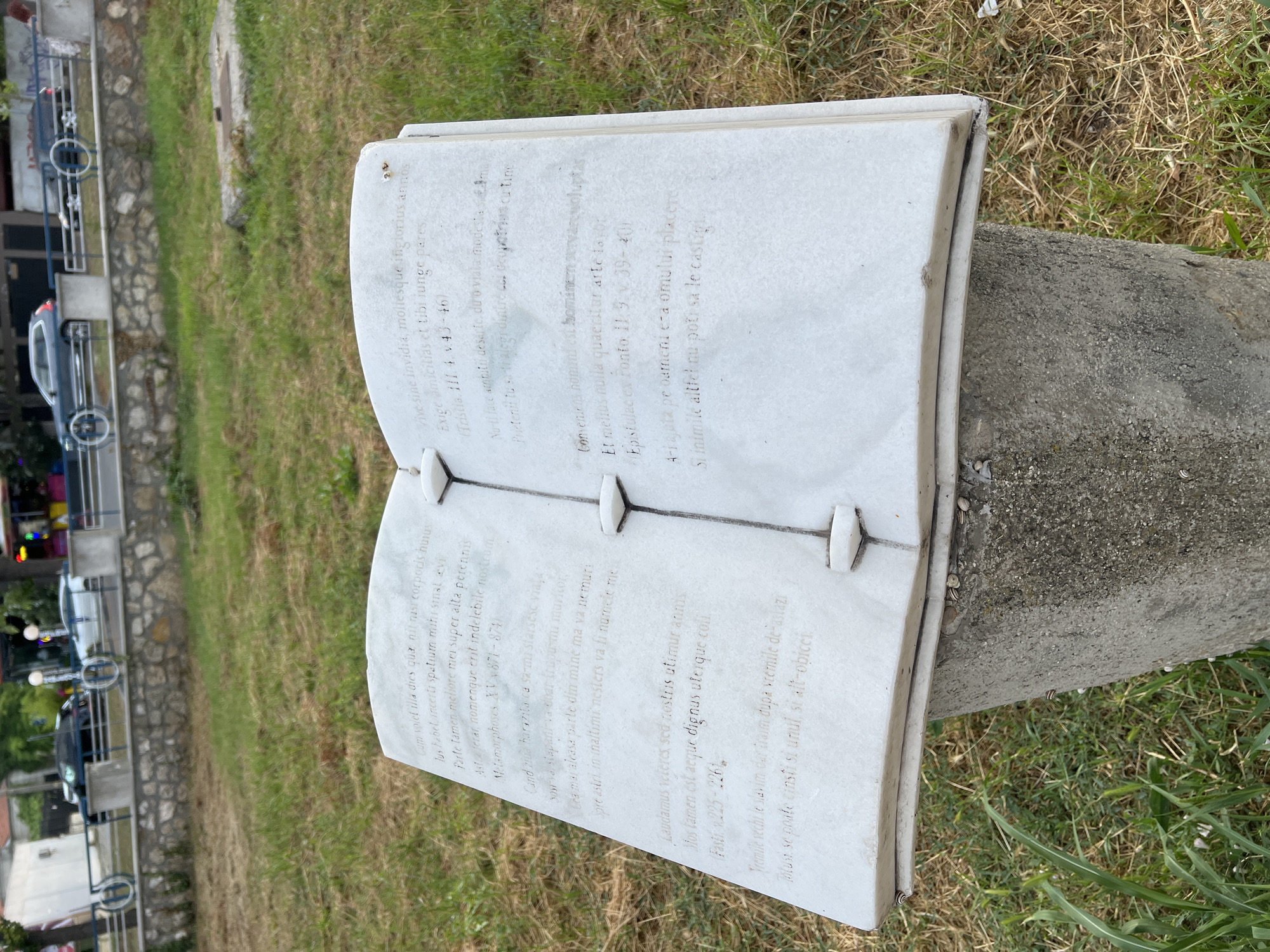 The image shows an outdoor sculpture of an open book made of white marble, resting on a concrete base. The book is inscribed with text in what appears to be Latin on the left page and Romanian on the right page. The sculpture is situated in a grassy area with some dry grass and a few scattered snails on the base. In the background, a road with cars and a stone wall can be seen, along with some buildings and greenery. The overall setting suggests a public space, possibly a park or a memorial site. The inscription on the left page is from Ovid's Metamorphoses, and the inscription on the right page is from a Romanian poem. This sculpture is located in Constanța, Romania, specifically in the park of the National Museum of History and Archaeology of Constanța.