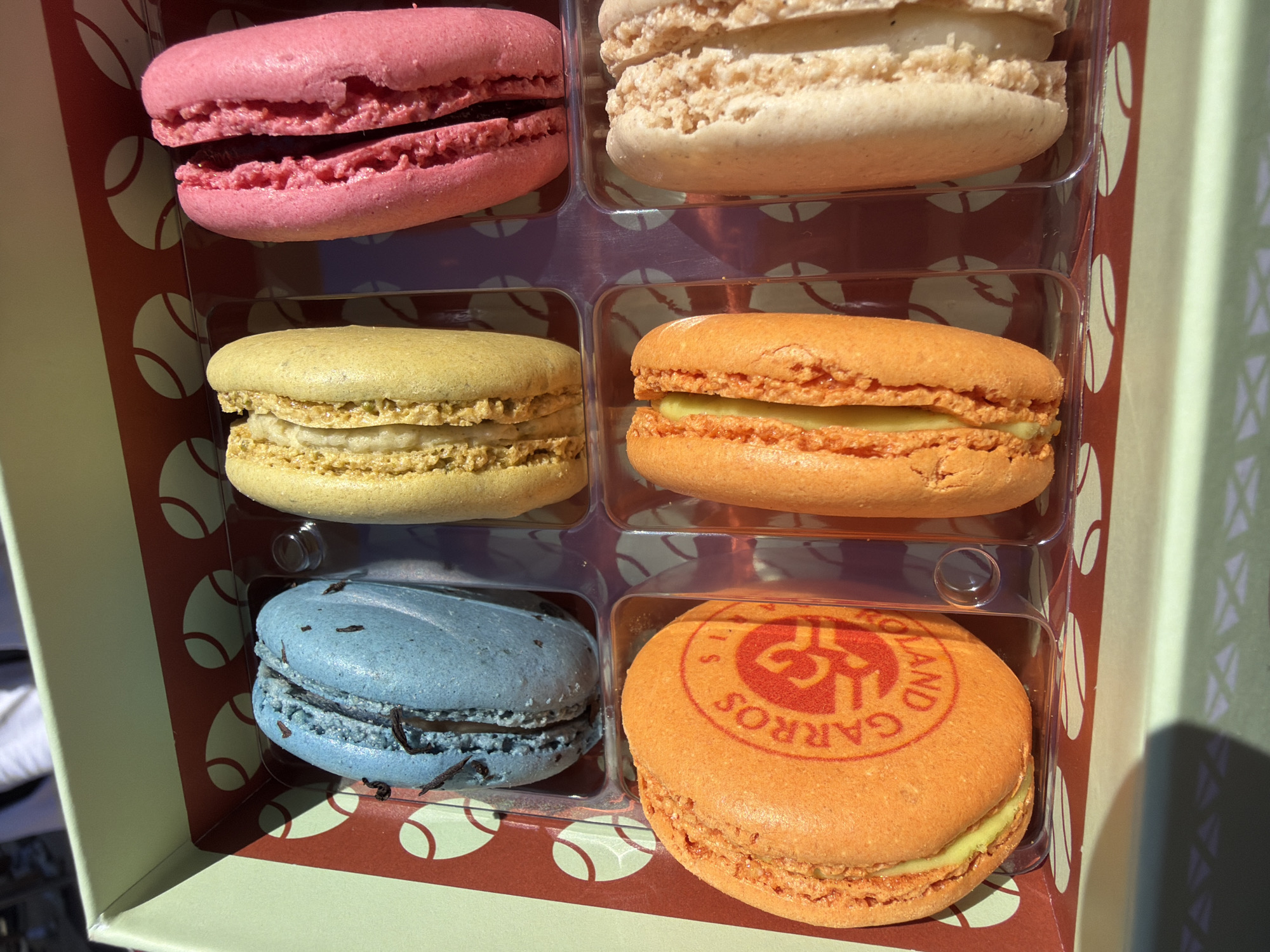 A close-up shot shows a box containing six macarons. The macarons are arranged in two rows of three, each in its own compartment. The macarons are different colors, including pink, beige, yellow, orange, and blue. The orange macaron in the bottom right corner has a logo that says "Roland Garros" on it, indicating a connection to the French Open tennis tournament held in Paris, France. The box has a pattern of tennis balls on its exterior.