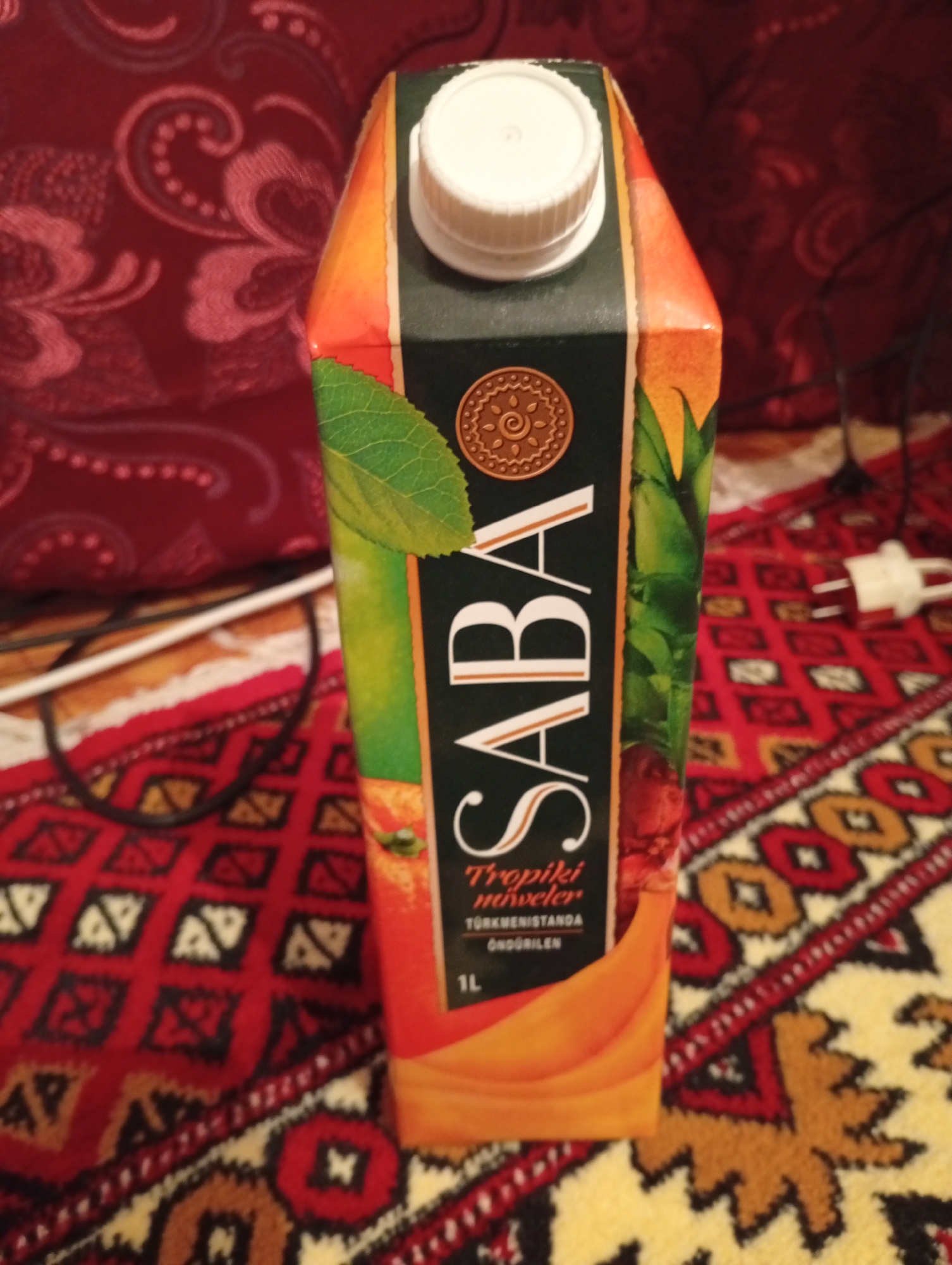 A juice carton is positioned vertically in the foreground, against a backdrop of a patterned red fabric and a red and yellow rug. The carton features the brand name "SABA" in large white letters. Below the brand name, smaller text indicates "Tropiki muwelewler" and "Türkmenistanda öndürilen," along with "1L." A white cap is visible at the top of the carton. The scene appears to be indoors, on a flat surface covered by a rug, with a red textile draped behind the carton. There are no people visible in the image. The image does not provide any explicit information about the city of Seýdi, Turkmenistan, beyond what is printed on the juice carton itself.