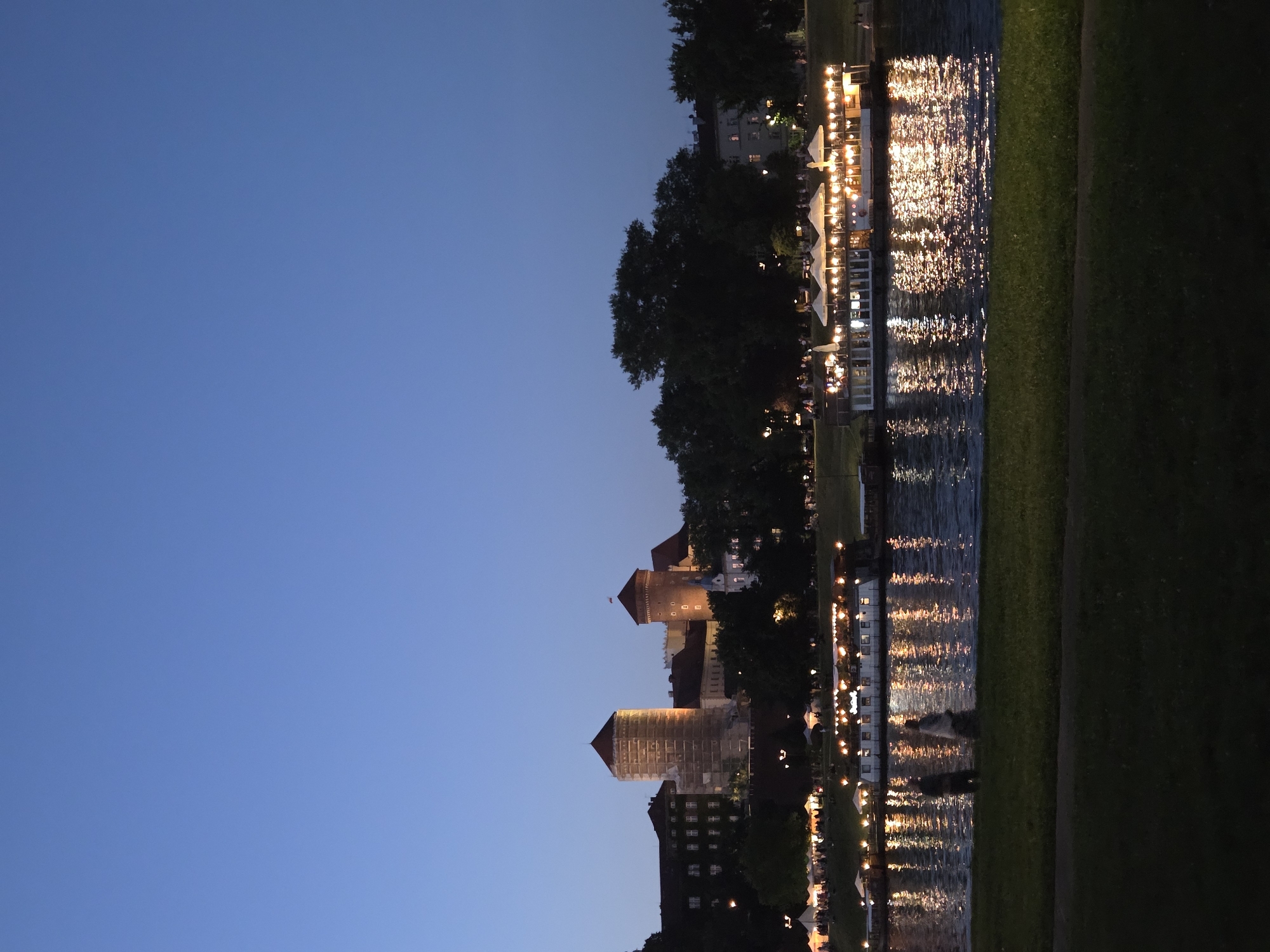 The image captures an evening view of the Vistula River in Krakow, Poland, with Wawel Castle and its surrounding buildings visible on the embankment. The sky is a deep, clear blue, transitioning into twilight. The castle features distinctive round towers, one of which is partially covered in scaffolding. Below the castle, a line of trees and buildings with illuminated windows can be seen.
Along the riverbank, there are several floating restaurants or boats, illuminated with numerous warm lights, their reflections shimmering on the dark water. White umbrellas are visible on the decks of these establishments, suggesting outdoor seating. Further down the riverbank, more lights indicate what appears to be a market or a gathering with tents.
In the foreground, a grassy embankment slopes down towards the river. Two figures, silhouetted against the illuminated water, are walking along the edge of the grass. The overall atmosphere is serene and picturesque, showcasing a lively evening scene in Krakow.