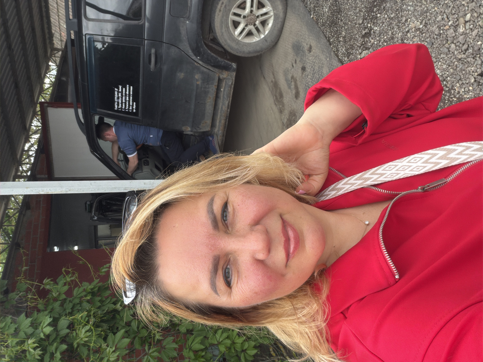The image is a selfie of a blonde woman in a red jacket, standing outdoors. She is smiling slightly and has her hand near her head. In the background, partially visible, is a dark SUV parked under a structure, possibly a garage or workshop. A man is working inside the SUV. There is text written on the SUV, presumably instructions or a warning. The location appears to be outdoors, possibly in Leningradskaya, Russia, based on the text on the vehicle. The ground is gravel. Some green vegetation is visible in the lower left corner.