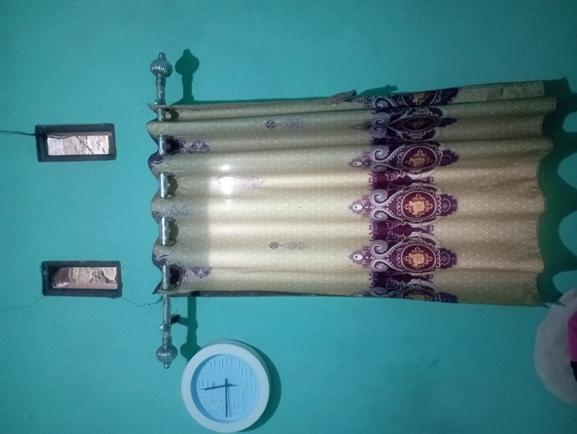 The image shows a teal-colored wall in a room. On the left side of the wall, there is a round, white analog clock. Above the clock, there are two small rectangular openings in the wall, resembling vents or small windows. In the center of the image, a beige curtain with a purple and gold ornate pattern hangs from a metal curtain rod. The curtain is pleated and covers a portion of the wall. To the bottom left, a blurry object, possibly a pillow or stuffed animal, is visible. The lighting in the room appears to be artificial, casting shadows from the curtain rod onto the wall. The overall impression is of a simple interior space. There are no people or animals clearly visible in the image. This scene could be found in a residential dwelling within the city of Maos Kidul, Indonesia, a region known for its diverse and often modestly furnished homes.