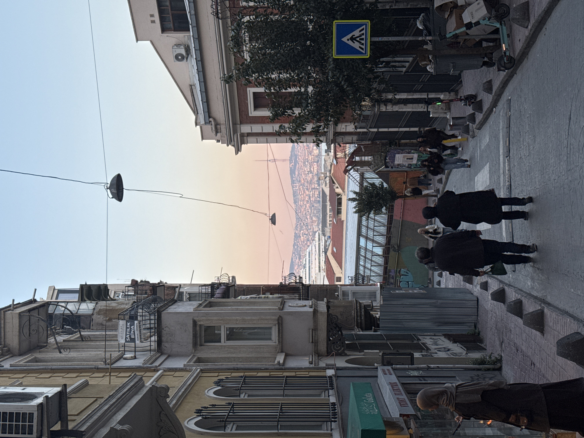 This image captures a narrow, sloping street scene in Beyoğlu, Turkey, likely during late afternoon or early evening as indicated by the gradient sky transitioning from light blue to a pale orange-pink hue in the distance. Multi-story buildings with varied architectural styles, some featuring balconies and intricate window bars, line both sides of the street.
On the left, a building with yellow facade details and ornate window grates is prominent. A single, dark street lamp is suspended from wires crisscrossing overhead. On the rooftops, a white sign reads "SATILIK BINA" (Building For Sale) and "FERHAT GERMEN".
The right side of the street features more buildings, one adorned with a prominent blue pedestrian crossing sign (a square with a white triangle and a walking person symbol) amidst tree branches. Several pedestrians are visible on the sidewalk and street; two individuals in dark clothing walk away from the viewer, and a small group is further down. Electric scooters, branded "BinBin", are parked on the sidewalk near trash bins and concrete bollards.
The street descends, revealing a panoramic view of a densely built city sprawling across hills in the background, partially silhouetted against the colorful sky. At the bottom left, storefront signs display "VERAY ELEKTRIK" (Veray Electric) and "ÇAĞLA". The scene depicts a typical urban moment, with daily pedestrian activity and visible city infrastructure.