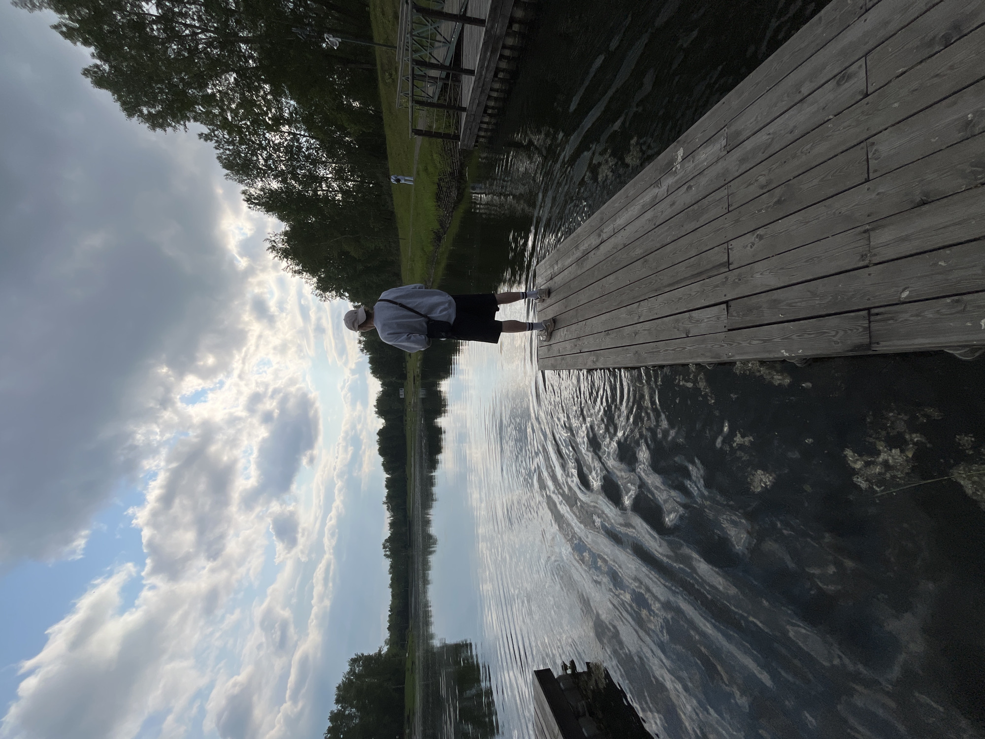 The image depicts a person standing on a wooden pier, facing a calm body of water. The pier is constructed of wooden planks and extends into the water. The person is wearing a light-colored sweatshirt, dark shorts, a baseball cap, and sneakers. They are holding a black bag slung across their body. The water is reflective, showing the sky and surrounding trees. The sky is partly cloudy, with a mix of blue sky and white clouds. Lush green trees line the far bank of the water, and a path or lawn area is visible in the distance, with another person walking on it. To the right of the main pier, there is another wooden structure that appears to be a dock or a walkway. The overall scene suggests a natural outdoor setting, possibly a park or recreational area by a lake or river. Based on the available information, the specific location within the city of Krasnoyarsk, Russia cannot be determined.