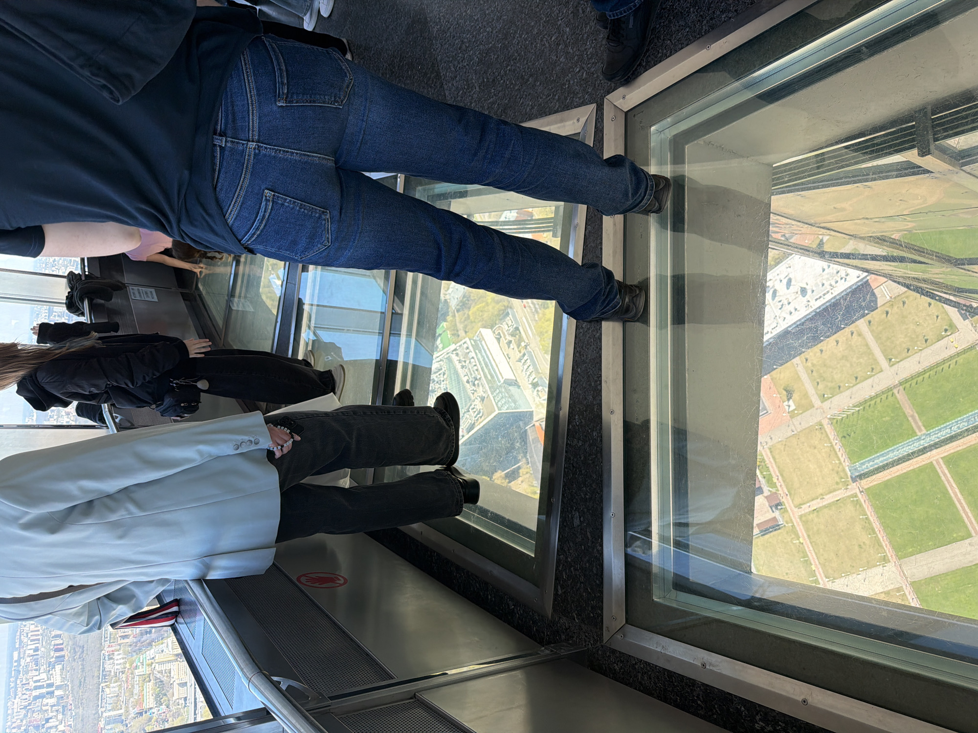 The image captures a view from a high vantage point, likely a skyscraper or tall building in Khimki, Russia, overlooking a cityscape and a park. People are standing on a glass platform, seemingly part of an observation deck. One person is wearing blue jeans and a dark top, standing on the glass floor with their legs stretched out. Another person is wearing a white blazer and dark pants, standing near a window with a view of the city. The reflection in the glass shows urban details, including buildings and green spaces. The park below is divided into rectangular sections, suggesting a formal garden or landscaped area. The environment is bright, with daylight illuminating the scene.