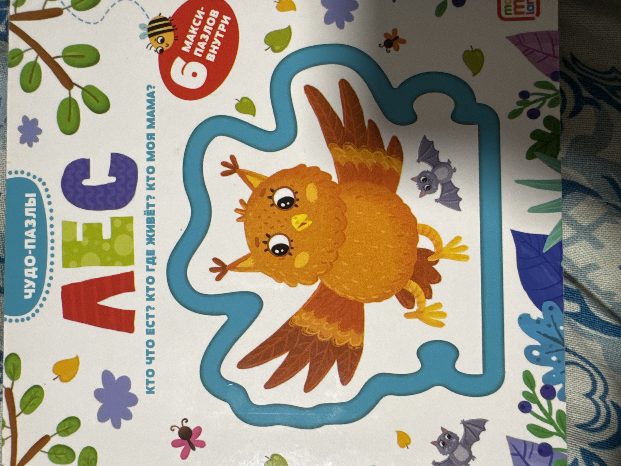 The image is a close-up, eye-level shot of the cover of a children's puzzle book. The book's title, "ЛЕС" (Forest), is prominently displayed in large, colorful letters across the top of the cover. Below the title, in smaller text, are the questions "КТО ЧТО ЕСТ? КТО ГДЕ ЖИВЁТ? КТО МОЯ МАМА?" (Who eats what? Who lives where? Who is my mom?). The cover features a cartoon illustration of an orange owl with large eyes and outstretched wings, set against a white background. Various cartoon forest elements, such as leaves, a butterfly, a bee, and two bats, are scattered around the owl. A blue, wavy border encloses the owl illustration. In the upper right corner, a red circle contains the number "6" and the Russian text "МАКСИ- ПАЗЛОВ ВНУТРИ" (MAXI-PUZZLES INSIDE). In the lower right corner, a logo with the letters "M" and "LAN" is visible. The bottom of the image shows a blurry pattern of fabric. The image does not contain any discernible elements that would allow for its localization to the city of Яромінскі сельскі Савет, Belarus.