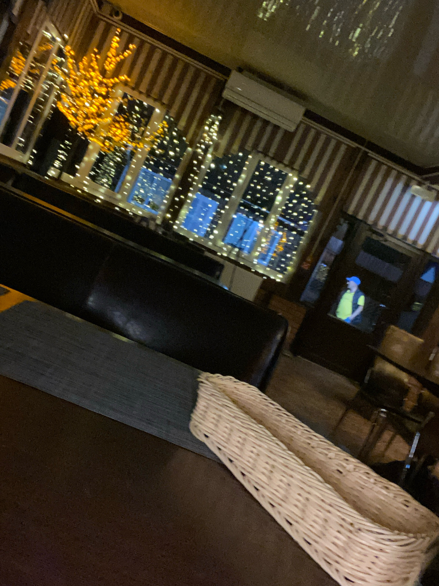 The image shows the interior of a dimly lit establishment, likely a cafe or restaurant, in Zhlobin, Belarus. The scene is dominated by dark furniture, including a large black sofa and a table in the foreground covered with a dark surface. A woven basket sits on the table. Large windows line one wall, adorned with numerous small, yellow string lights that create a festive atmosphere. Outside the windows, the lights of other buildings are visible, suggesting an urban setting. In the background, a doorway reveals a person wearing a yellow vest and a blue cap, possibly an employee. The walls are decorated with vertical striped wallpaper, and an air conditioning unit is mounted high on one wall. The overall impression is one of a relaxed, possibly evening, setting.