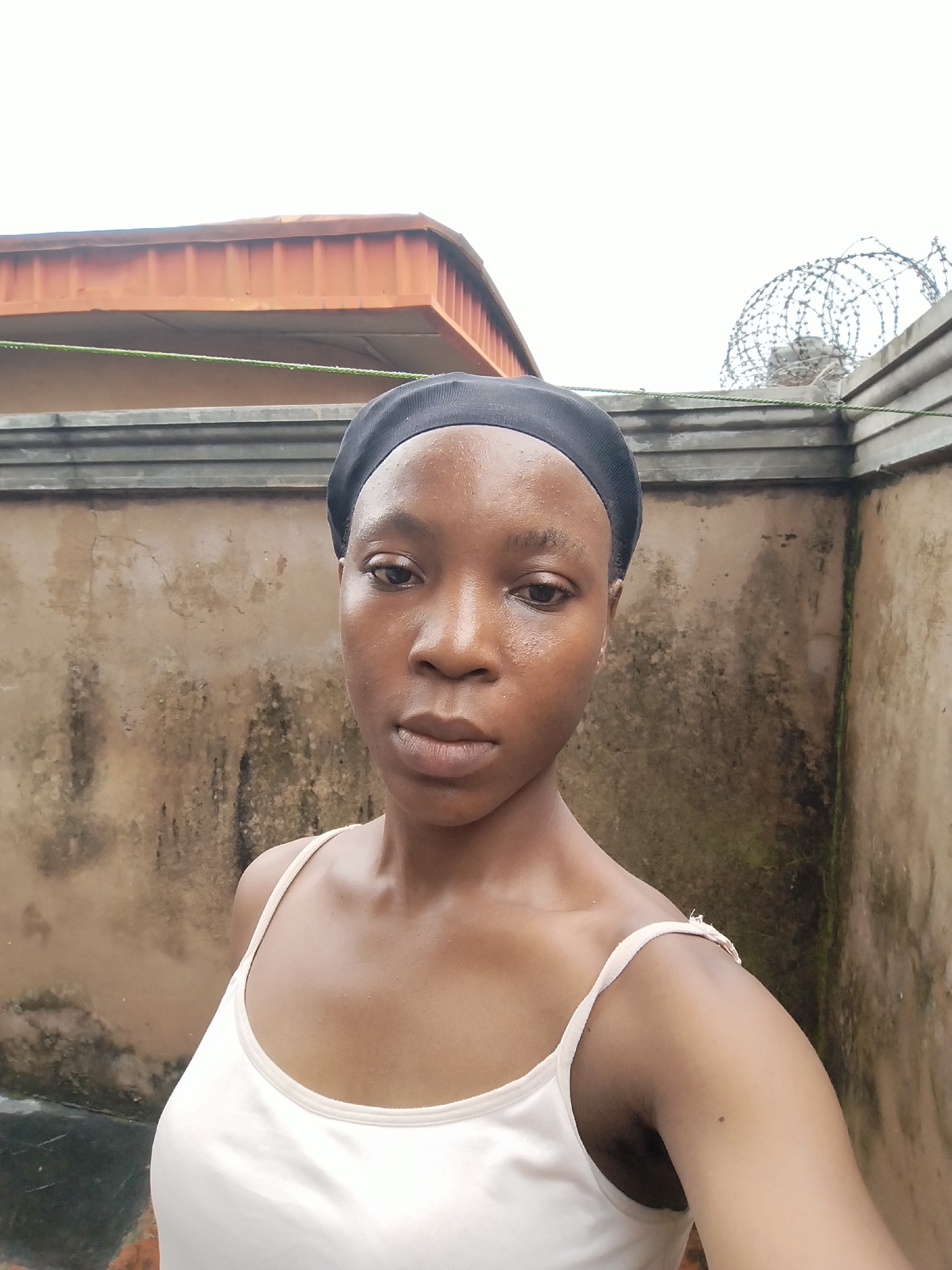 A young woman, wearing a black head covering and a white tank top, poses for a selfie outdoors. She has dark skin and her face is turned towards the camera. Behind her, a textured, dirty wall extends across the frame. Above the wall, parts of a reddish-brown roof and a gray concrete structure with barbed wire are visible against a bright, overcast sky. The environment appears to be a residential area in Isiehor, Nigeria.