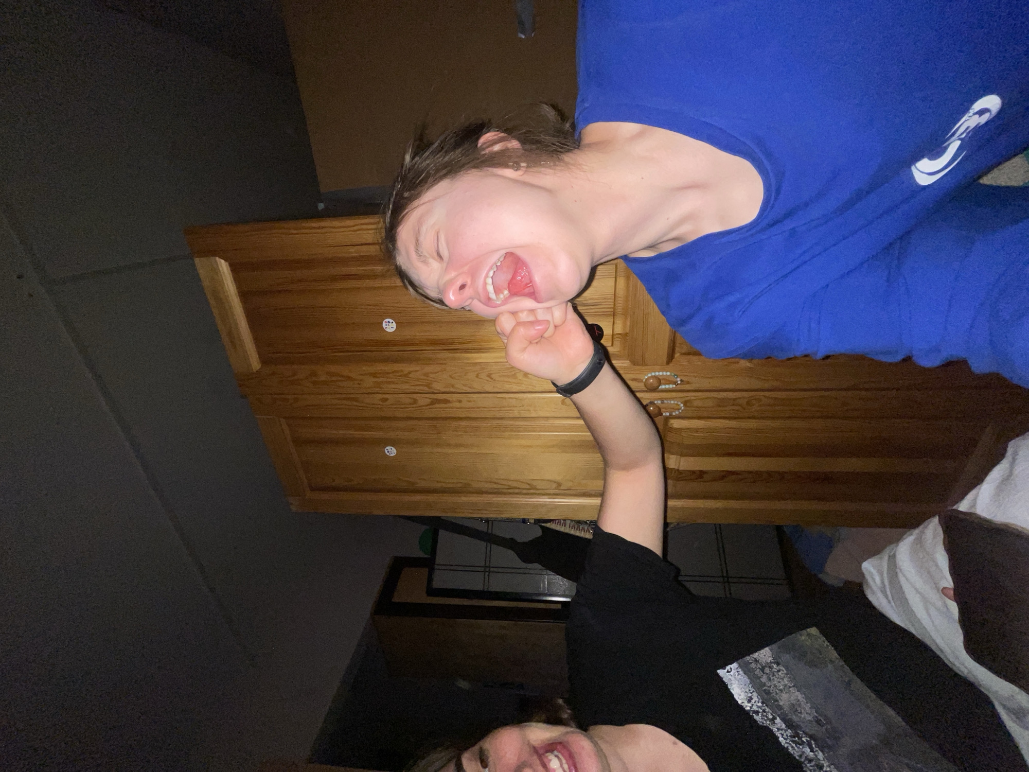 Two people are visible in the image, positioned with a dresser in the background. The person at the top, wearing a blue shirt, is contorting their face with their hand. The second person, wearing a black shirt, has one arm raised. The setting is an indoor space. The location is not identifiable as Moscow, Russia.