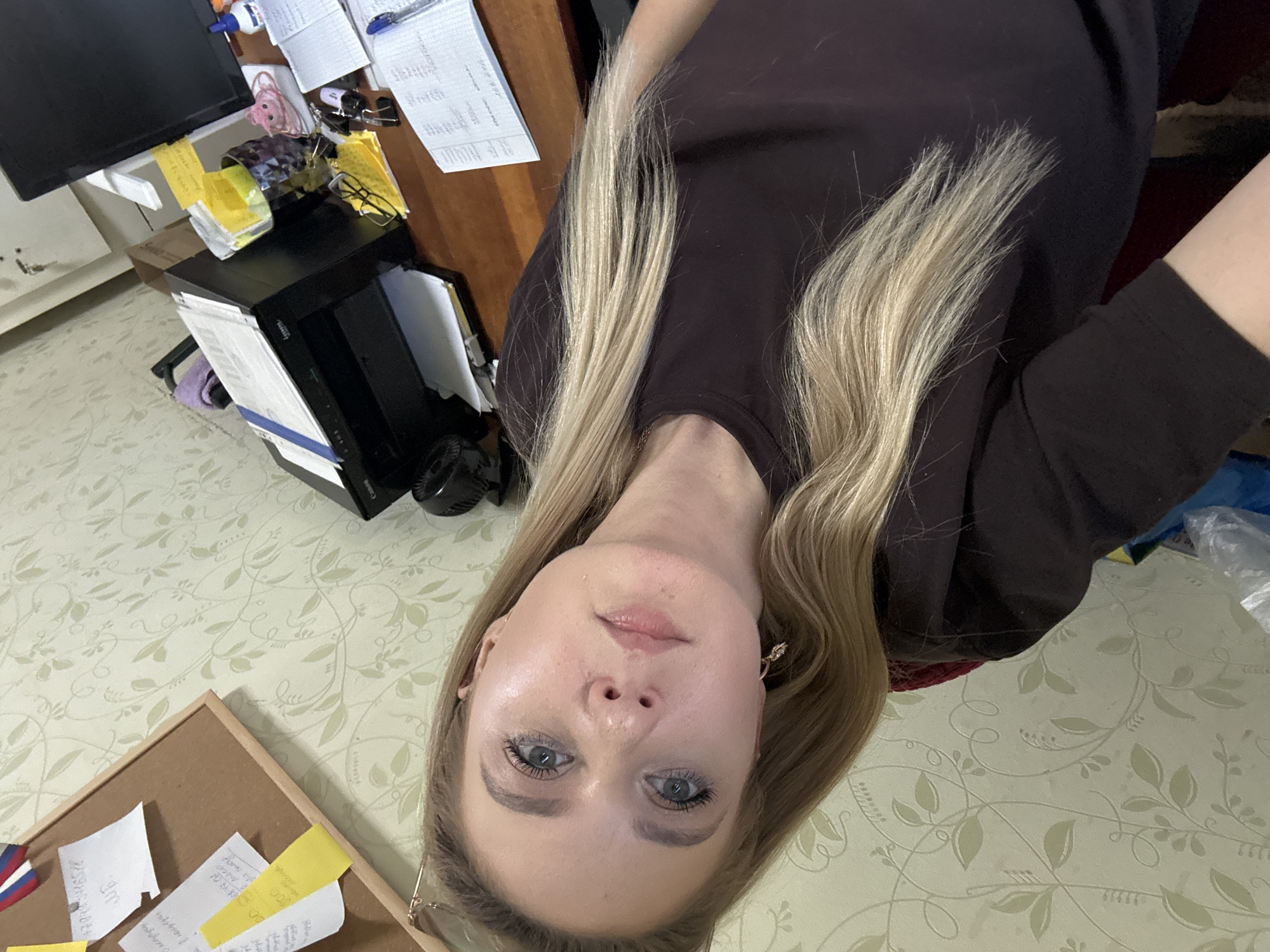 The media file shows an upside-down, close-up, selfie-style shot of a woman with blonde hair and blue eyes. She is wearing a dark brown shirt. Her makeup is visible, including mascara and eyeshadow. She is wearing earrings. The background appears to be an office or study, with a desk, computer monitor, papers, and a corkboard visible. The lighting suggests it could be daytime. The woman's expression is neutral. Some papers in the background have handwritten text, but it is not legible.