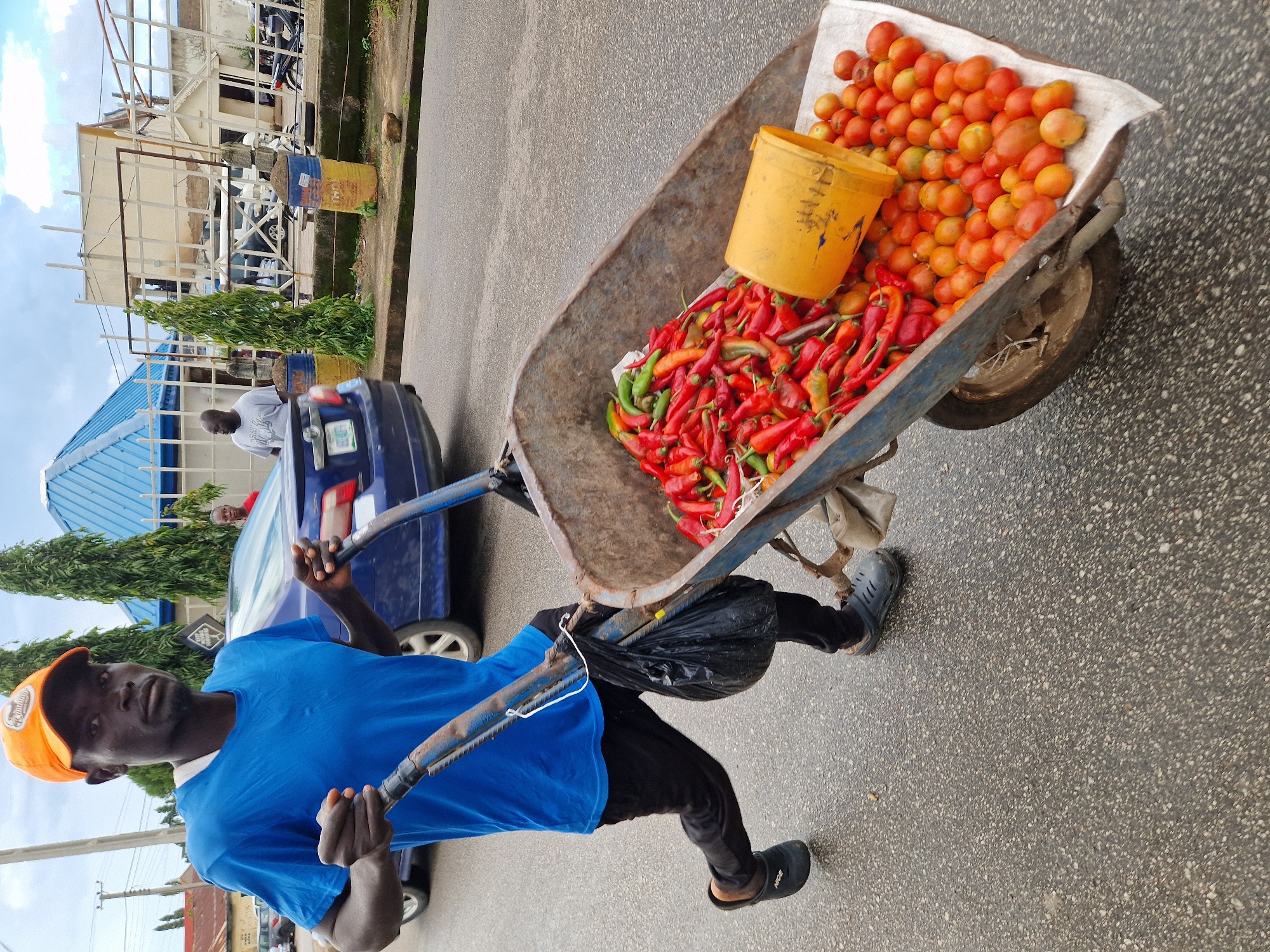 A dark-skinned man wearing a blue t-shirt, black pants, and an orange cap is pushing a wheelbarrow filled with red and green chilies and tomatoes on an asphalt road. He is facing the camera and looking directly at it. The wheelbarrow is old and rusty, with a yellow bucket lying on its side on top of the tomatoes. In the background, a blue car and several buildings can be seen. The sky is cloudy. The scene appears to be in an urban environment.