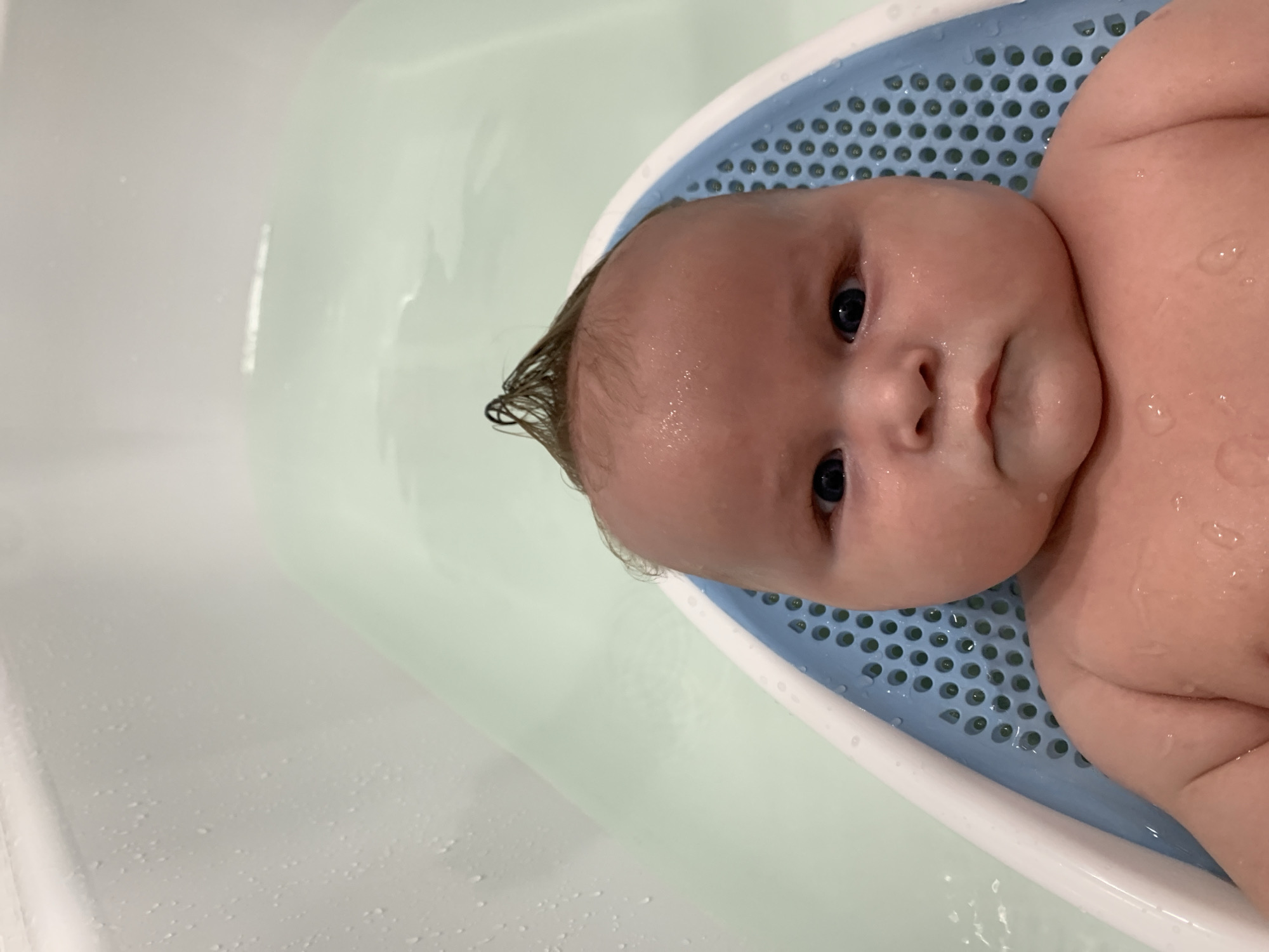A close-up, overhead, waist-up shot shows a chubby infant with dark blue eyes and a small tuft of dark hair styled like a mohawk, sitting in a baby bathtub. The baby's skin is wet and glistening with water droplets. The bathtub is light green with a blue mesh insert. The background is a blurred white bathtub wall with water droplets. There is no indication of the city of Stavropol, Russia, or any specific location within a city.