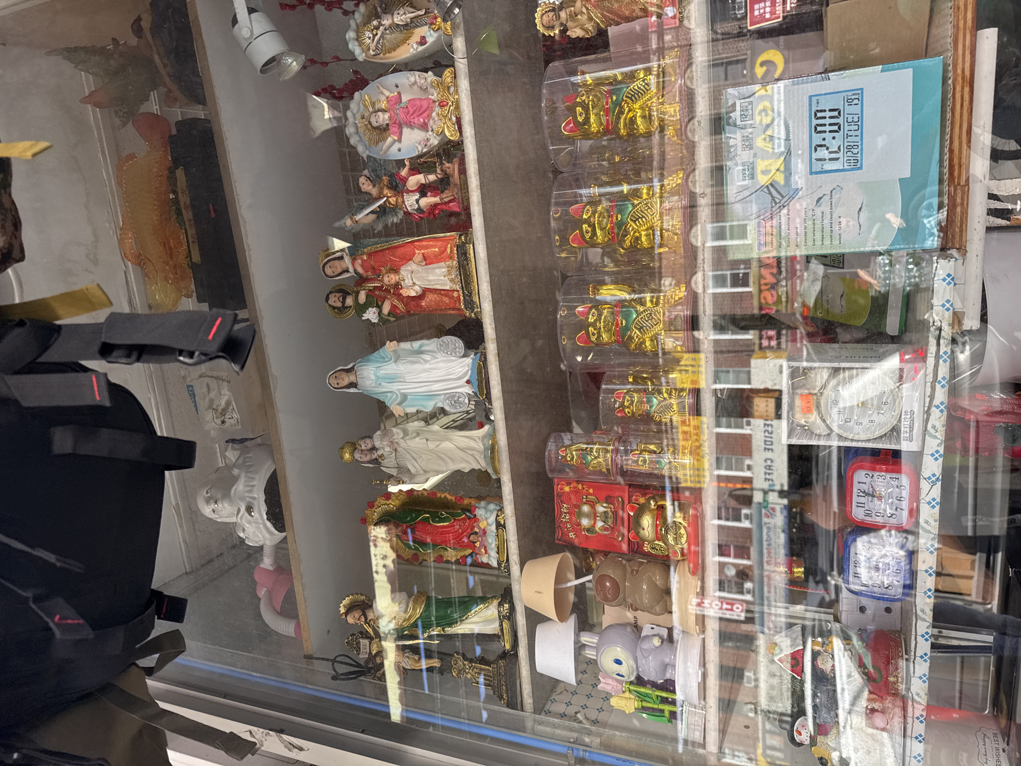 The image shows a display shelf in what appears to be a store window. The objects on display include religious statues of Jesus, Mary, and an angel, as well as decorative items like lucky cats, a smiling rabbit figure, and two small lamps. Further down, there are several clocks, including one digital clock displaying "12:00" and the date "10/28 TUE 19". The overall environment suggests a retail setting, possibly a gift shop or an antique store, located in New York City, United States of America. The merchandise is arranged on multiple shelves within a glass enclosure, reflecting the surroundings.