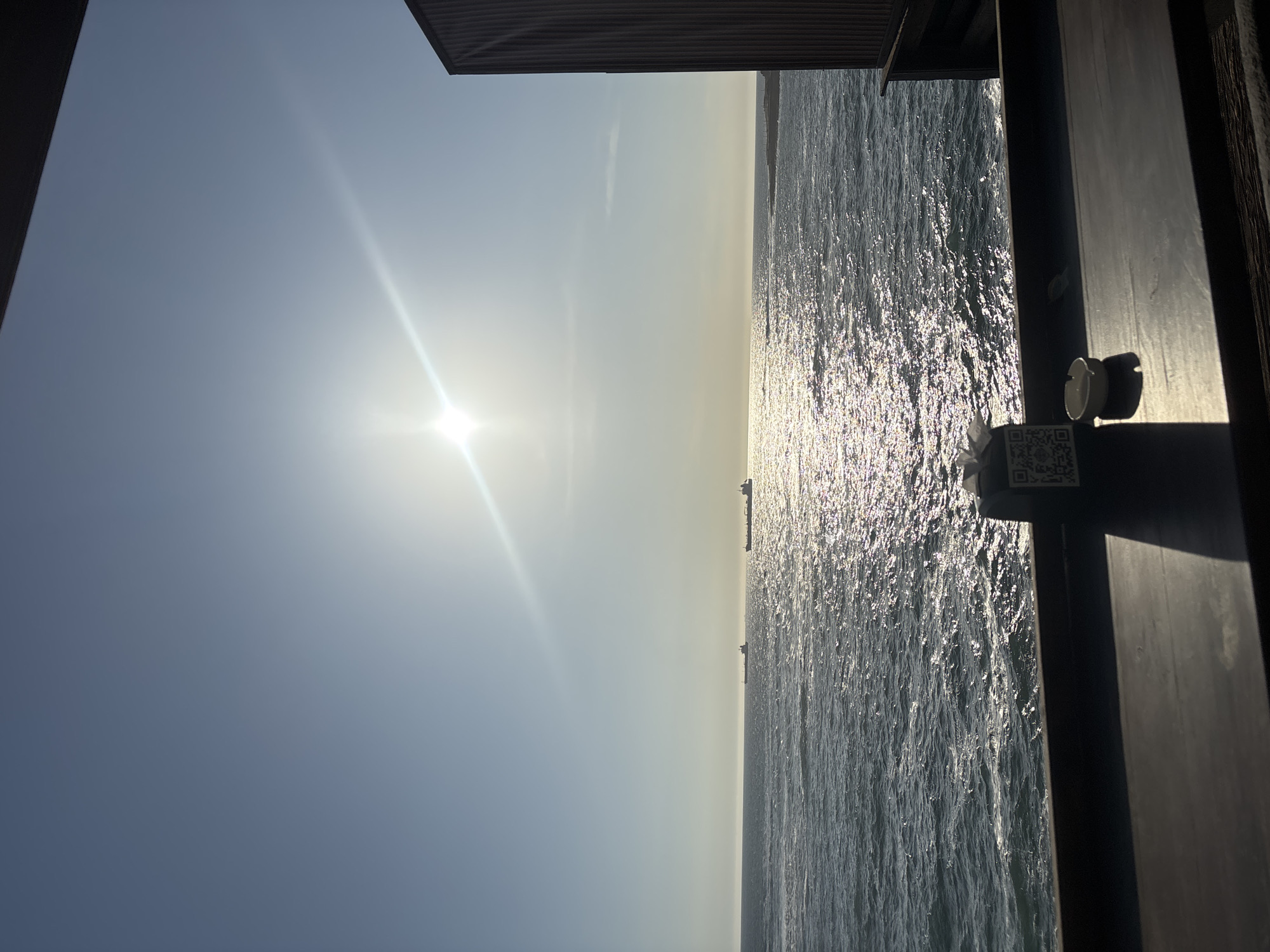 The image shows a view of the Pacific Ocean from Arica, Chile, on a bright, sunny day. The sun is high in the sky, creating a glare on the water's surface. Two ships are visible on the horizon. The shot appears to be taken from a building or structure, with parts of the building visible in the frame, including a security camera and a QR code sticker attached to a surface.