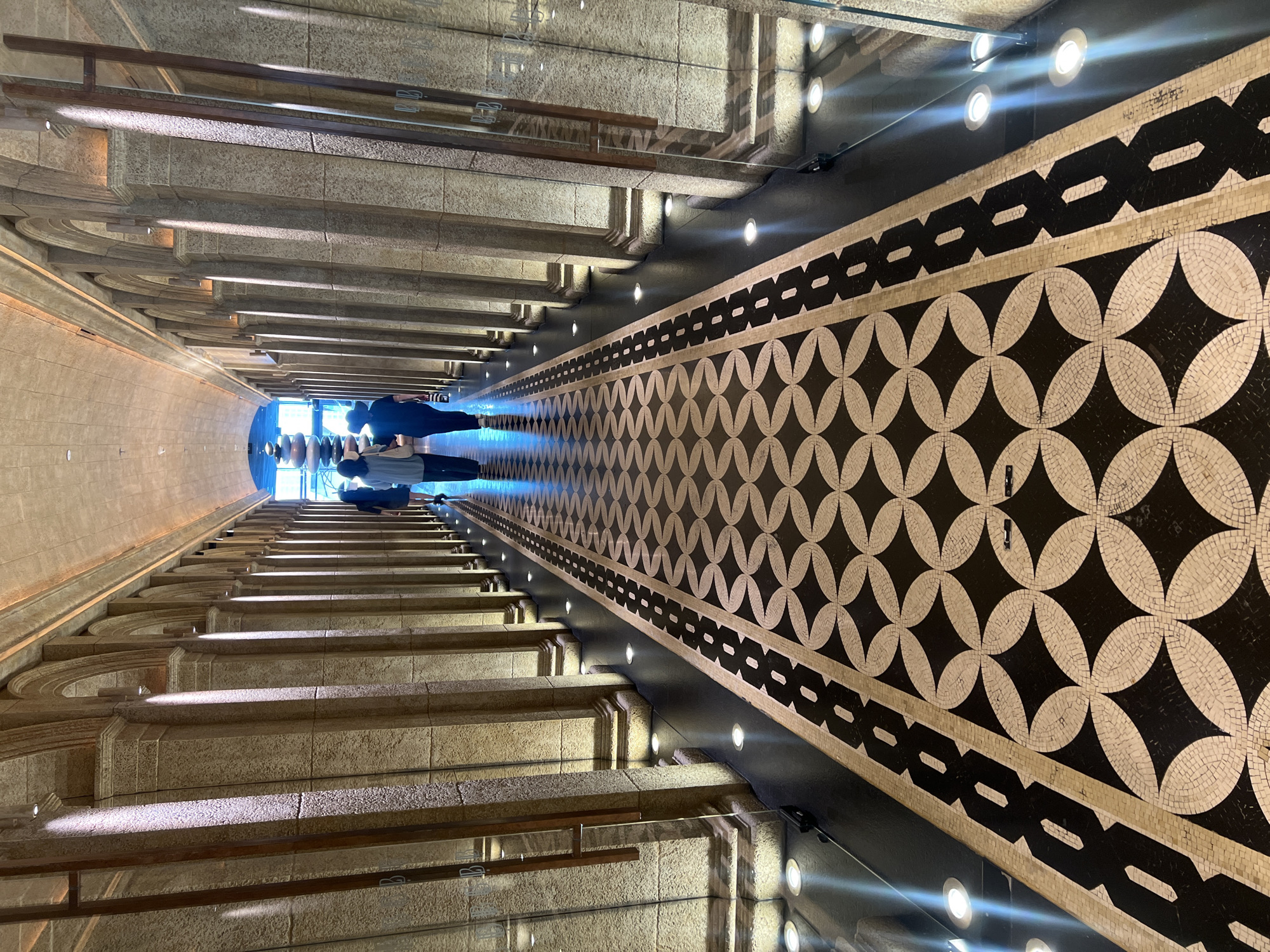 The image displays a grand indoor architectural space, captured from a tilted, upward-looking perspective along a long, ascending corridor or staircase. The scene is characterized by strong architectural lines and a largely monochromatic color palette.
On the left, a series of substantial, light-colored stone steps or ledges ascend, flanked by robust stone walls featuring classical architectural details and subtle arches. Small, recessed spotlights are integrated into the lower sections of these steps, casting distinct patterns of light and shadow. A dark handrail parallels the incline.
The right side presents a striking black and white mosaic pattern on a vertical surface running alongside the path. The mosaic features a repeating geometric motif of interlocking circles and squares, creating an intricate visual texture. Above this mosaic, a line of bright, circular recessed lights illuminates the area. A transparent glass railing, with metallic fixtures, separates the mosaic wall from the central ascent, reflecting some of the light.
At the far, elevated end of the corridor, two human figures are visible. They appear as dark silhouettes against a strong, bright light source, which emits a blueish glow into that section of the space, suggesting a possible large window or exit.
The overall impression is one of a sophisticated and imposing interior, indicative of a public building, museum, or luxury establishment. The strong geometry, interplay of light and shadow, and classical design elements contribute to a sense of depth and grandeur within this setting in Dubai, UAE. No discernible text is visible.