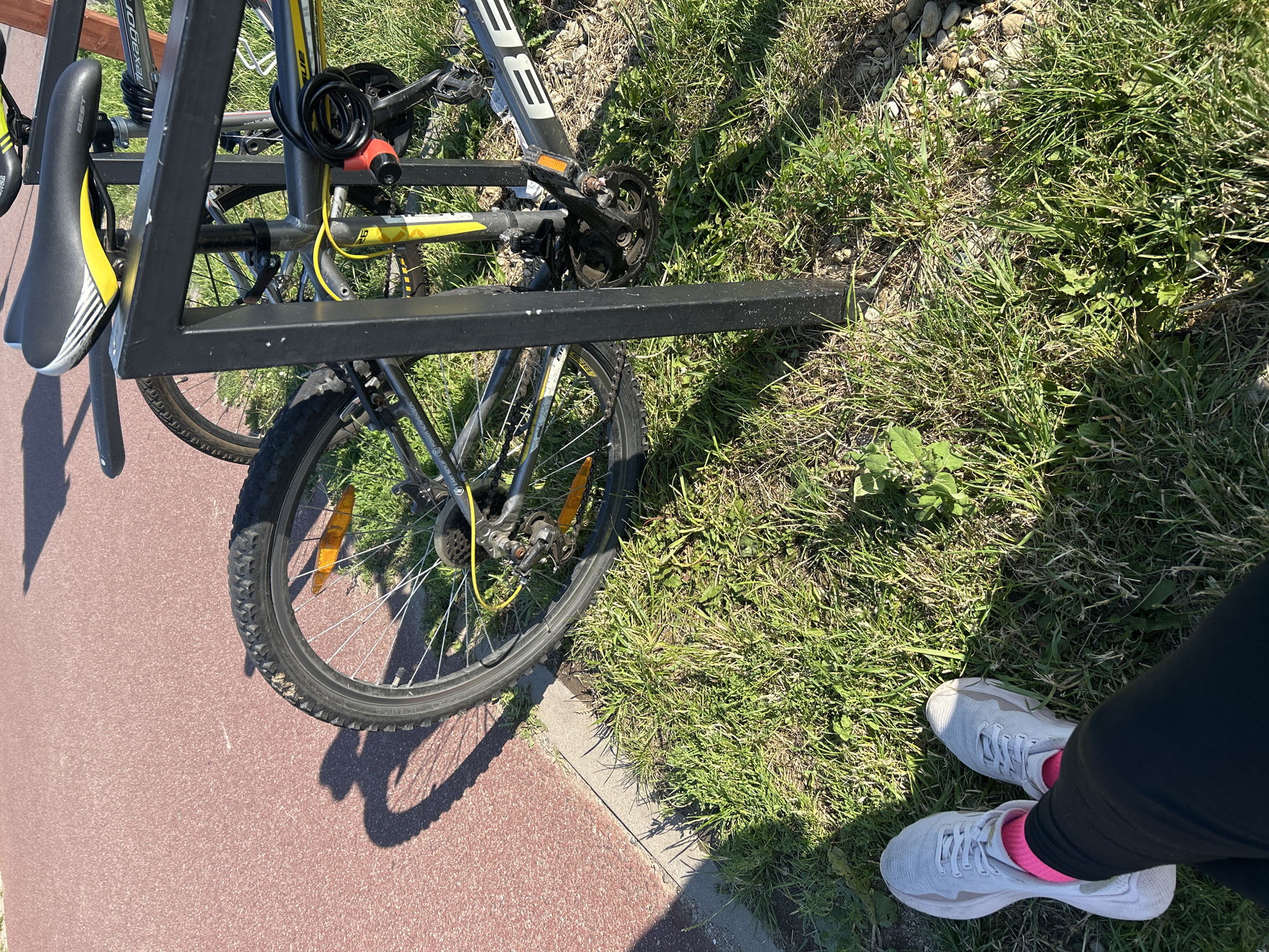 An outdoor scene shows two bicycles parked at a bike rack in Bałdowo, Poland. The bicycles are silver with yellow accents. One bicycle has a black seat with yellow stripes. Both bicycles are secured with a red and black cable lock. The ground is covered in green grass and a red path runs along the left side of the image. In the foreground, a person's legs are visible, wearing white sneakers and pink socks. The person appears to be sitting or resting. The sun is casting shadows across the scene.