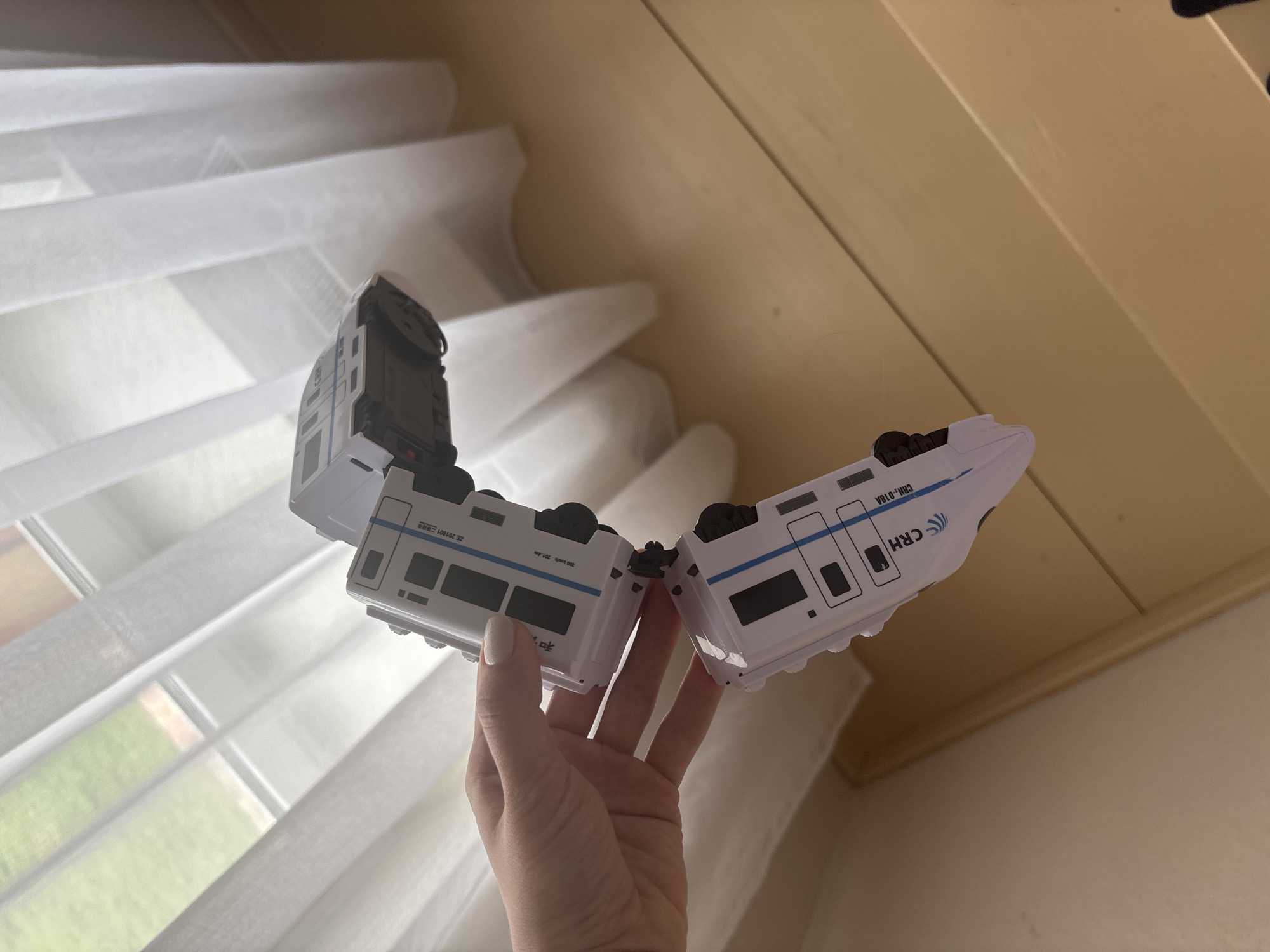 A photograph shows a Caucasian hand holding two halves of a white and blue toy train against a background of a drawn window shade. The train cars are separated at their connecting point. The toy train has details of Chinese text and symbols on its sides. The image seems to be taken indoors, likely at or near Ферма, Russia. The exact location is unknown.