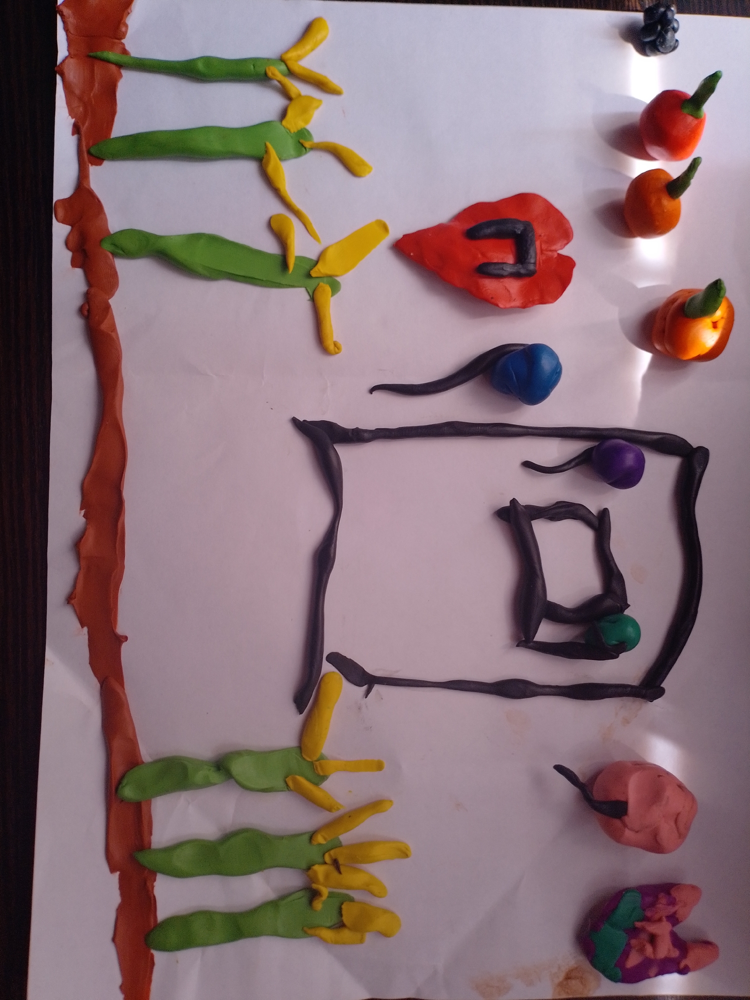 The image shows a variety of colorful clay sculptures arranged on a white sheet of paper. In the upper left quadrant, there are three green, bean-shaped objects with yellow, finger-like protrusions extending from them, positioned vertically. To the right of these, there are several small, round, red and orange objects with green stems, resembling miniature fruits or vegetables. A red, leaf-like shape with a black U-shaped element is also present. In the central portion of the image, a black, square-like frame encloses a white space, with dark blue, purple, and green spheres placed around and within it, connected by thin, black, wavy clay strands. Towards the bottom left, another set of three green, bean-shaped objects with yellow, finger-like extensions is visible. The lower right quadrant contains a pink, lumpy object with a small black strand and a multi-colored object composed of purple, green, and pink clay. The background is a white surface, with a dark wooden surface visible on the far left edge of the frame. There is no identifiable location in the city of Tashla, Russia, within this image.