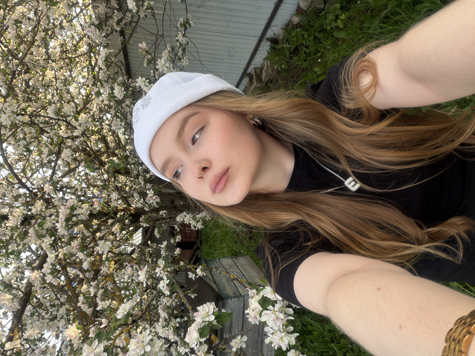 A young woman takes a selfie. She is wearing a white beanie and a black t-shirt. She has long, brown hair and a silver necklace with a square pendant. Behind her is a tree with white flowers, a wooden shed, and green grass. The photograph was taken in the city of Кадряково, Russia.