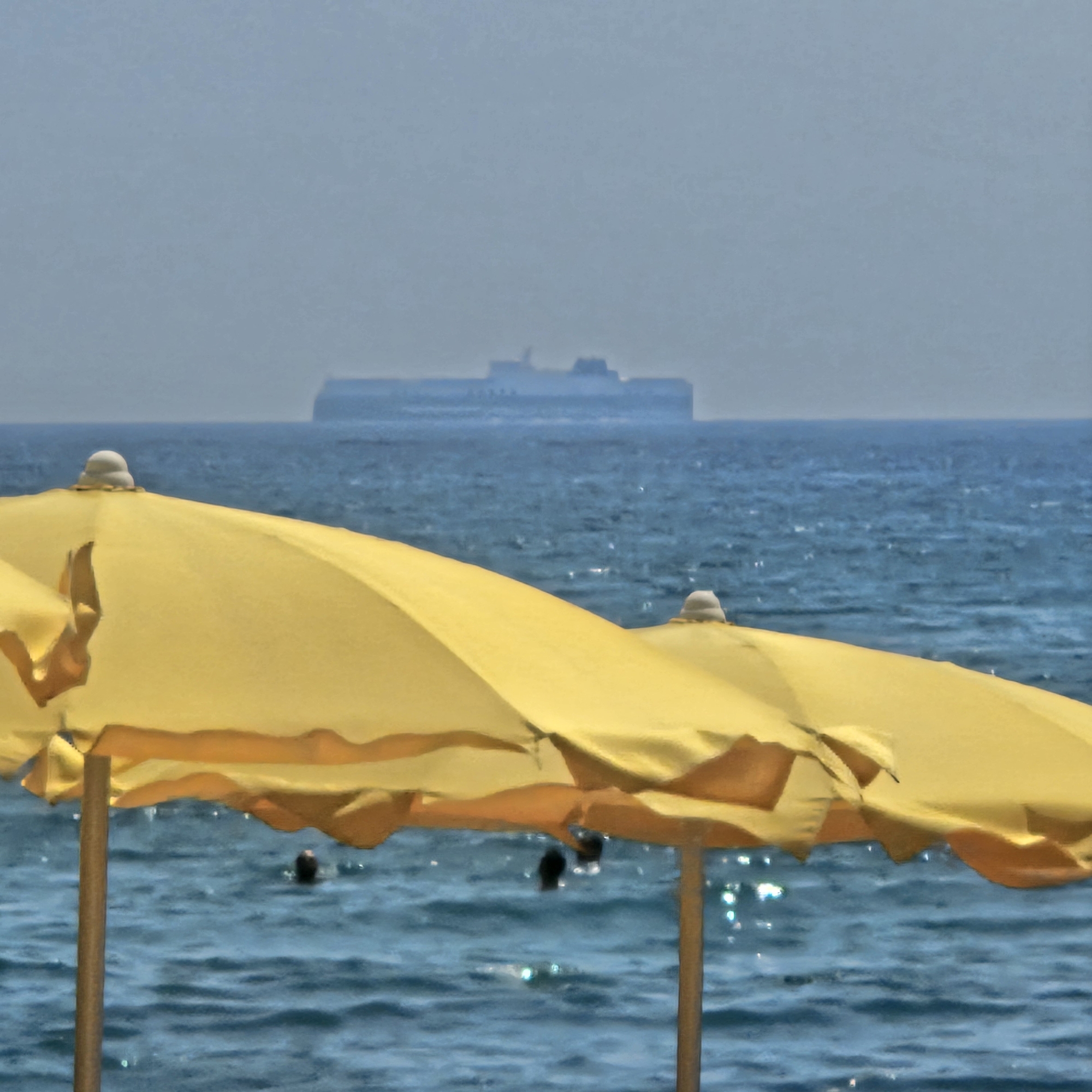The image depicts a scene at a beach in Savona, Italy, with a focus on yellow beach umbrellas in the foreground. The umbrellas are partially obscuring the view of the sea and the distant horizon. The sea is a deep blue, with gentle waves reflecting the sunlight. In the far distance, a large, light-colored ship is visible on the horizon, appearing somewhat hazy due to the atmospheric conditions. The sky is a pale blue-gray, suggesting it might be a slightly overcast or hazy day. A few people are visible in the water, swimming or wading, their heads just above the surface. The overall impression is a serene beach scene, with elements of both relaxation and activity.