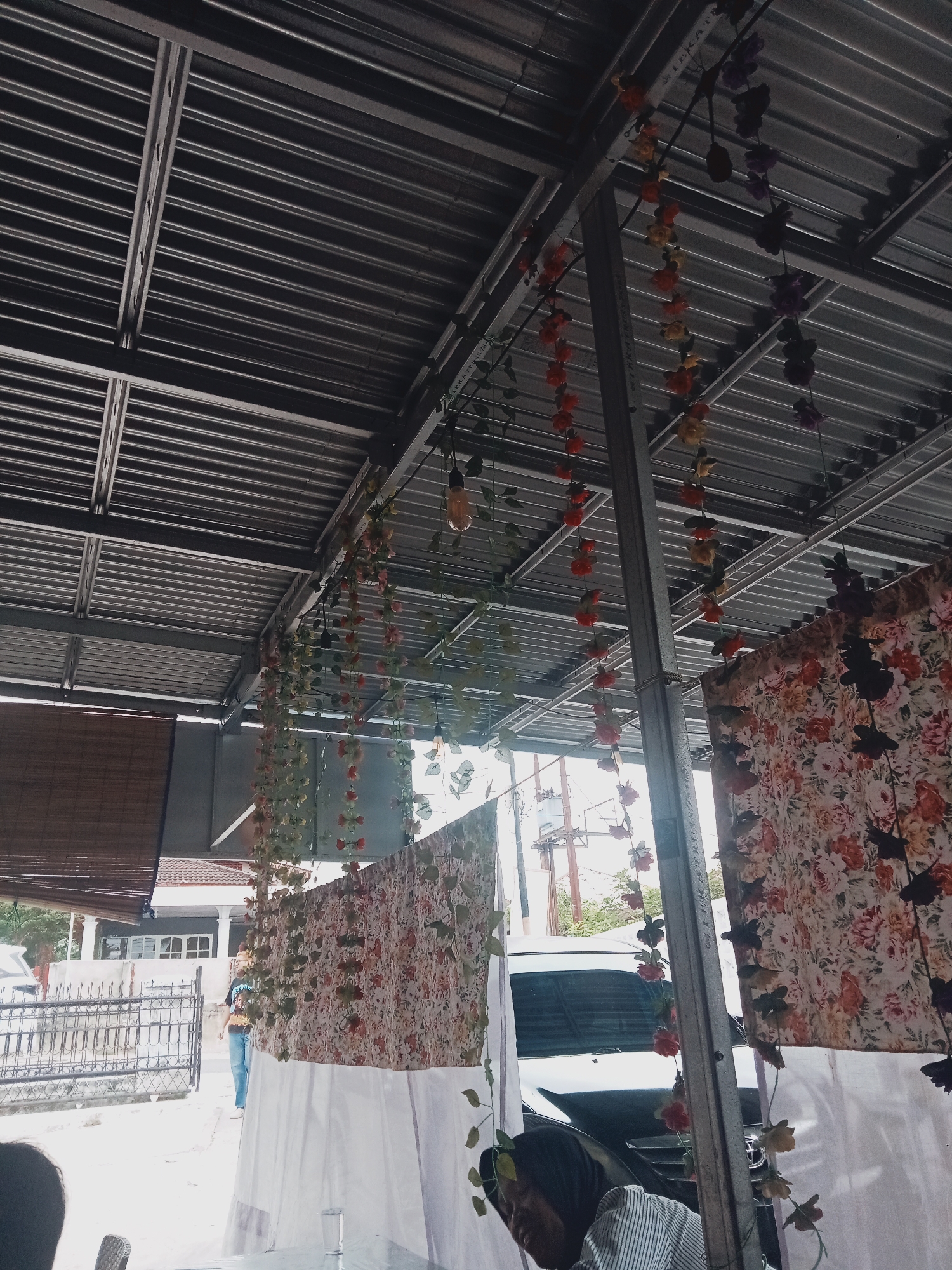 The image depicts an outdoor seating area under a corrugated metal roof, likely at a food stall or restaurant in the city of Balik Alam, Indonesia. Floral garlands with red, yellow, and purple flowers hang from the roof structure, adding a decorative element. A patterned fabric, featuring a floral design with red, pink, and cream colors, is draped to the right. White fabric is also visible, possibly as part of the stall's setup. In the background, a person in jeans and a t-shirt walks past a metal fence and a building with white pillars. A white vehicle is parked behind the floral fabric. The overall impression is of a casual, possibly open-air dining or resting space.
