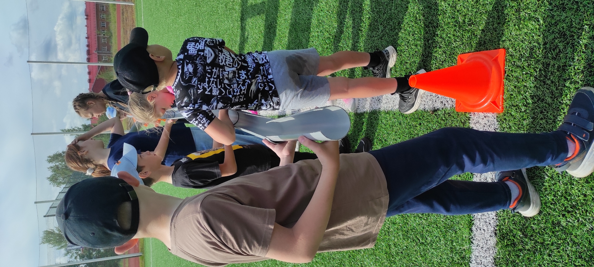 The image depicts a group of children and young adults gathered on a bright green artificial turf field, likely a sports field, under a partly cloudy sky. In the foreground, a young boy wearing a black baseball cap and a brown t-shirt is visible from the waist up, holding a long gray cylindrical object. To his right, another boy in a black t-shirt with a yellow design and a light blue cap looks ahead. Next to him, a younger child with blonde hair leans down, partially obscured. Further to the right, a boy in a black t-shirt with a white graphic print and gray shorts faces away from the camera. Two young women stand in the background, one with dark braided hair and a black t-shirt, the other with her hand on her forehead and wearing a dark blue t-shirt. The field is marked with white lines, and an orange traffic cone is placed on the ground near the right edge of the frame. In the far background, a red-roofed building and trees are visible beyond a metal fence. The overall scene suggests an outdoor activity or event taking place in a suburban or semi-rural setting, potentially a school or community sports ground. Based on the provided metadata, the location is the city of Noviy, Russia.