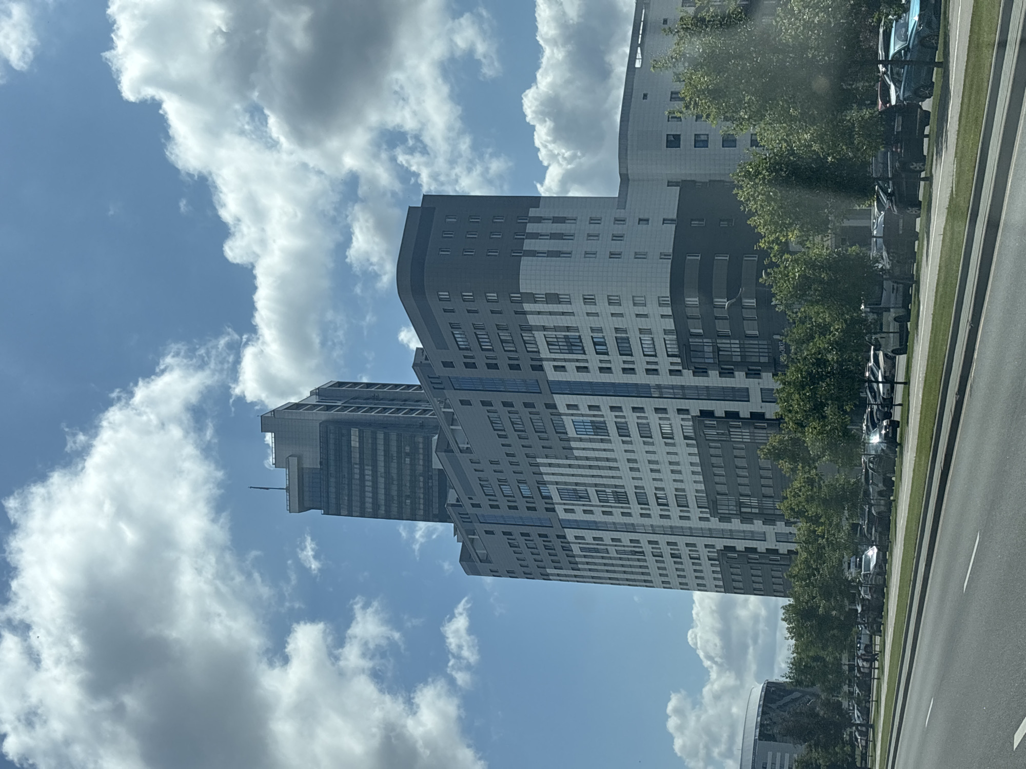 The image shows a modern residential complex in Minsk, Belarus, on a partly cloudy day. The complex features two prominent buildings: a taller, slender skyscraper with a darker facade and a more expansive, multi-story building with a lighter, patterned exterior. The buildings are characterized by their clean lines, numerous windows, and varied architectural elements, including rounded sections and contrasting color blocks.
In front of the residential complex, a tree-lined street stretches along a wide, asphalt road. Cars are parked along the side of the road and on a green median strip. The trees are lush and green, indicating it is likely summer. The sky is blue with scattered white clouds, some of which are large and fluffy, suggesting fair weather with some atmospheric movement.
To the left of the frame, another modern building with a curved facade is partially visible, suggesting an urban development area. The overall impression is one of contemporary urban living in a well-maintained city.