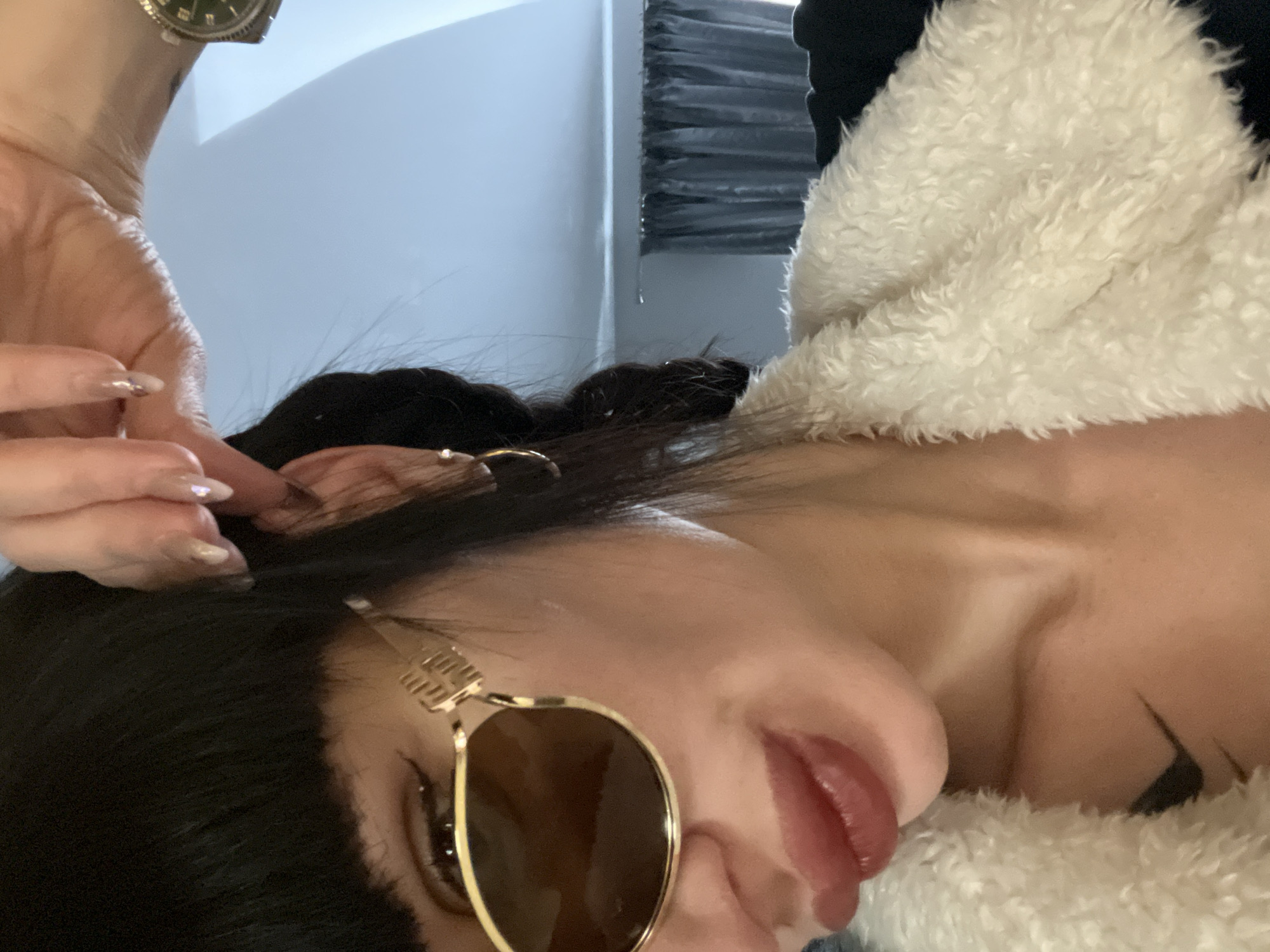 A person with dark hair, light skin, and sunglasses is close to the camera. The person is wearing a fluffy, white jacket. A hand with manicured nails is near the person's head. The location is not discernible from the provided image.