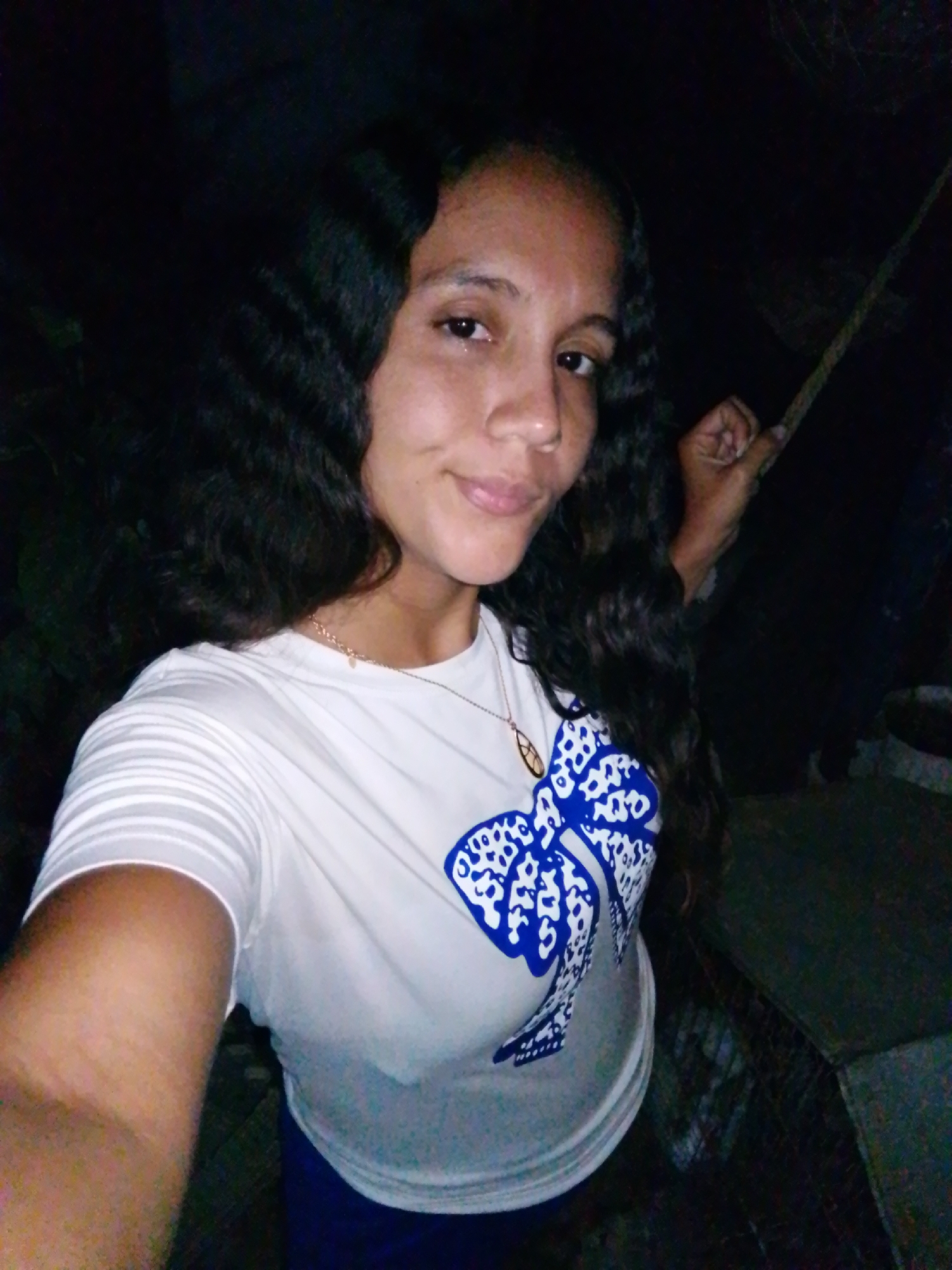 A woman with long, dark curly hair stands against a dark, indistinct background. She wears a white t-shirt with a blue and white patterned design and a thin gold necklace. She is holding a thin, light-colored object (possibly a stick or rope) in her right hand. The location is unknown, so I can't provide information on Juan Griego, Venezuela.