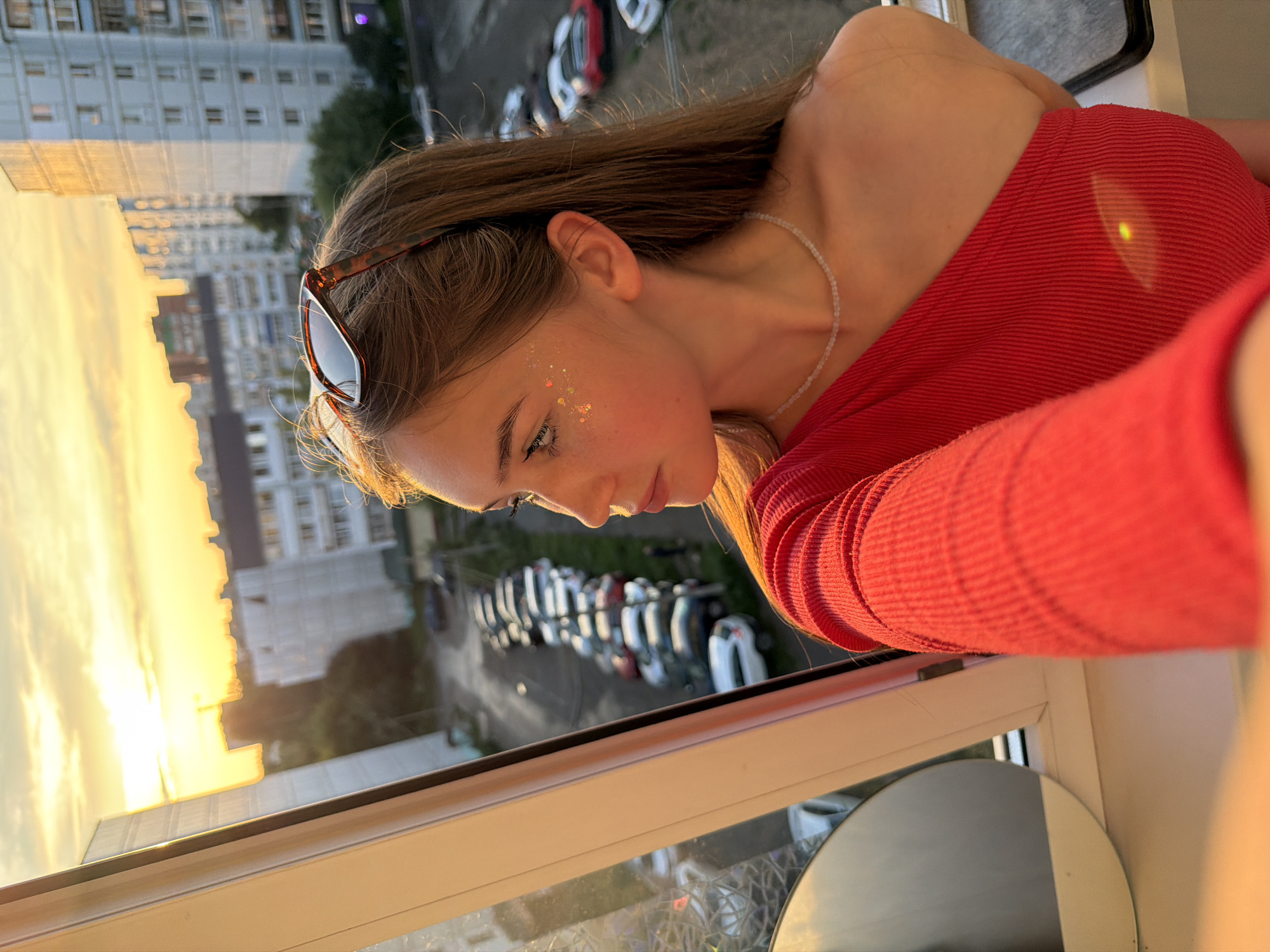 A young woman with long brown hair is leaning out of a window, looking to the left. She is wearing a red one-shoulder top and has glitter on her face. Sunglasses are perched on her head. In the background, a cityscape is visible at sunset, with buildings and a street lined with parked cars. The scene appears to be in Chaikovsky, Russia, a city known for its industrial development and proximity to the Kama River.