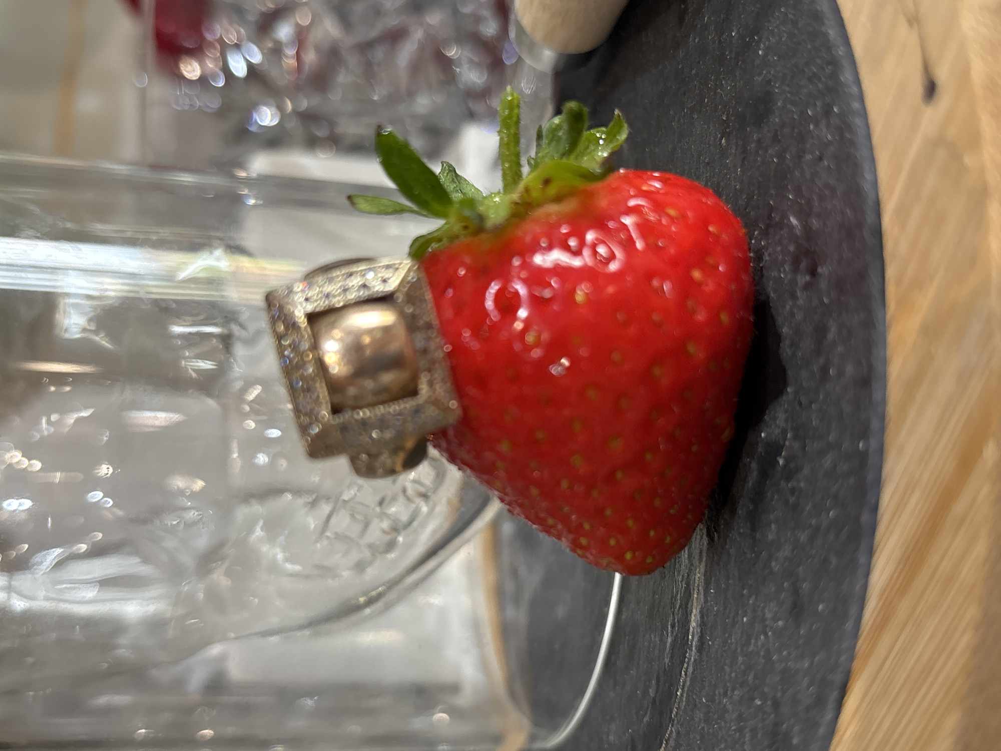 The image shows a close-up, angled shot of a single strawberry resting on a dark, textured surface, possibly slate, which is next to a wooden surface with vertical grain. The strawberry is vibrant red with visible seeds and a green stem with leaves. A ring, encrusted with small, sparkling stones and featuring a larger square centerpiece, is placed around the base of the strawberry, partially obscuring it. The background is blurred, revealing a clear glass object, possibly a jar or bottle, with light reflecting off its surface, creating bokeh effects. Hints of red objects are visible in the further background, also blurred. The lighting appears bright, with strong highlights on the strawberry and the ring, suggesting an indoor setting, possibly a kitchen or dining area. No text is visible in the image. The overall mood is still and somewhat luxurious, given the presence of the jeweled ring. The setting is not explicitly detailed, but the wooden element could suggest a rustic or natural aesthetic alongside the polished ring and fresh fruit.