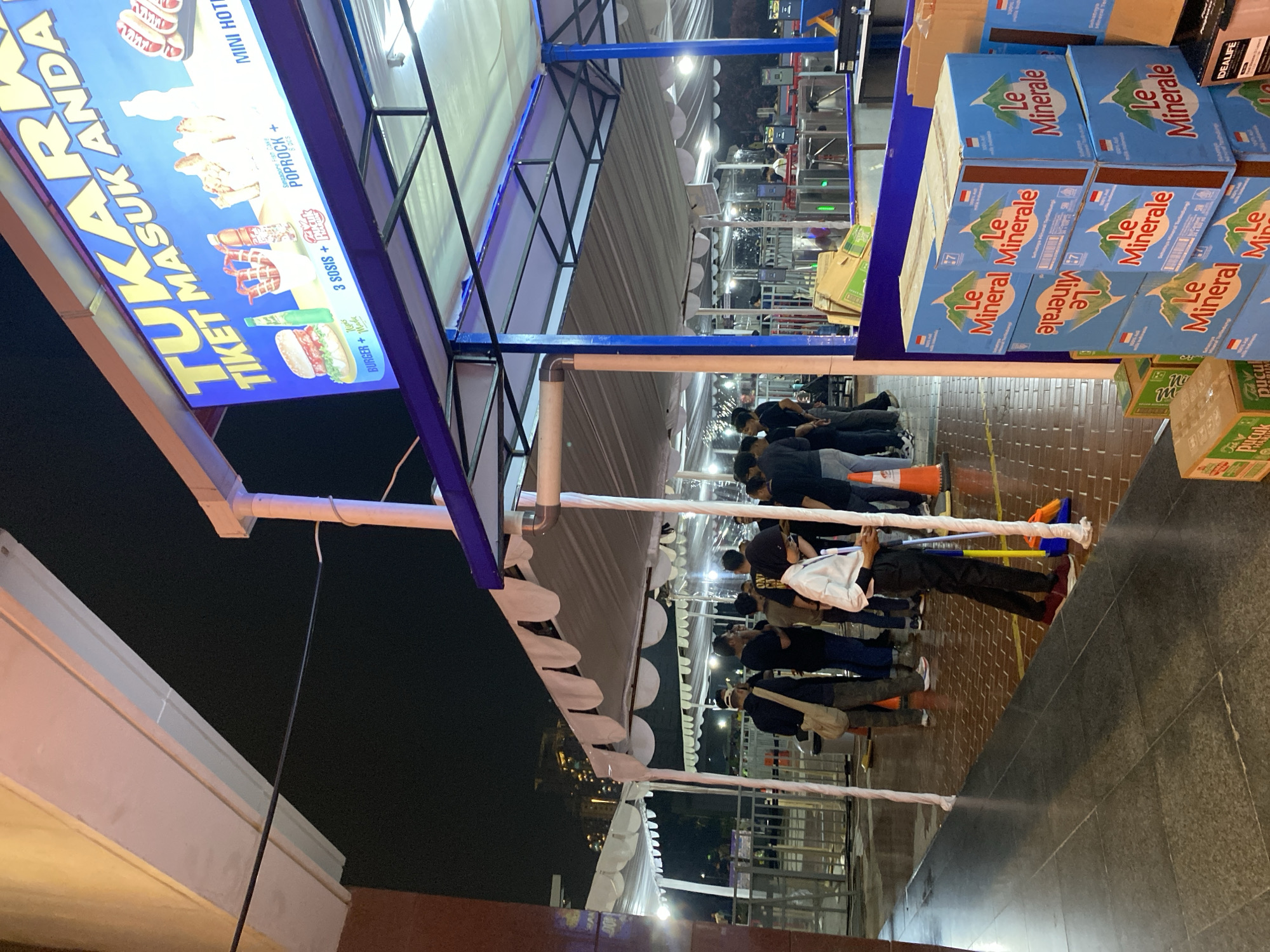 A group of people stand in front of a food stall under a tent at night. The stall has a large sign advertising "TUKARKAN TIKET MASUK ANDA" (Exchange Your Entrance Ticket) and various food items. Several boxes of "Le Minerale" bottled water are stacked in the foreground. In the background, there are turnstiles for entry and exit. The scene is lit by artificial lighting.