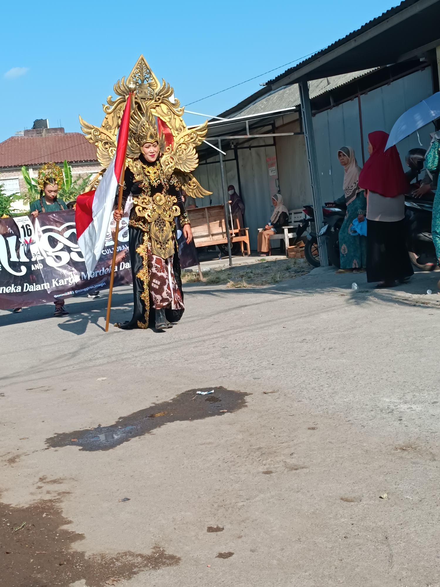 In the city of Imogiri, Indonesia, on a paved road under a clear blue sky, a parade participant walks forward, holding a wooden staff with a vertically oriented Indonesian flag. The individual is adorned in an elaborate, traditional costume featuring intricate gold embellishments on a black base, a winged headdress, and decorative leg coverings. To the left of the flag bearer, another participant, wearing a similar headdress and a green tunic, holds a dark banner with white text. In the background, a building with a corrugated metal roof is visible, and several onlookers, some wearing hijabs, stand to the right, with one person holding a light blue umbrella. The road surface is worn, with water stains and debris.