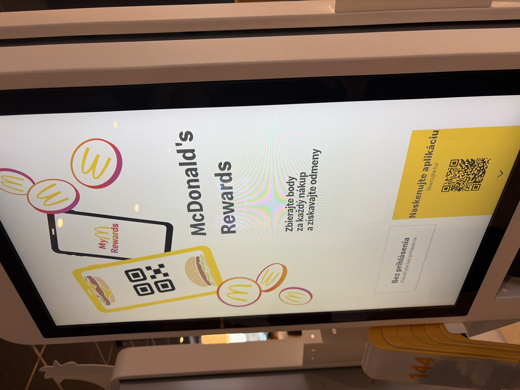 The image shows a close-up of a digital kiosk screen, likely located in a McDonald's restaurant. The screen displays information about McDonald's Rewards, with graphics of a phone showing a QR code and a burger. Text in Slovak is present, including "MyM Rewards," "Zbierajte body za každý nákup a získavajte odmeny" (Collect points for every purchase and earn rewards), "Bez prihlásenia Pokračujte bez prihlásenia" (Without logging in Continue without logging in), and "Naskenujte aplikáciu Sklenujte tu!" (Scan the app Scan here!) accompanied by a QR code.
The kiosk appears to be a self-service ordering or information terminal. The overall lighting suggests an indoor environment with some ambient light. There are no people visible directly interacting with the kiosk in the frame, though the context implies a public space. The background is out of focus, but hints of the restaurant interior, including yellow-colored elements possibly related to the McDonald's branding, are visible. A sign with the number "144" is partially visible on the left side of the frame, possibly indicating a queue or section number. Based on the Slovak language displayed on the screen, the location is in Slovakia. Without further contextual information, the specific location within the city of Nitra cannot be determined.
