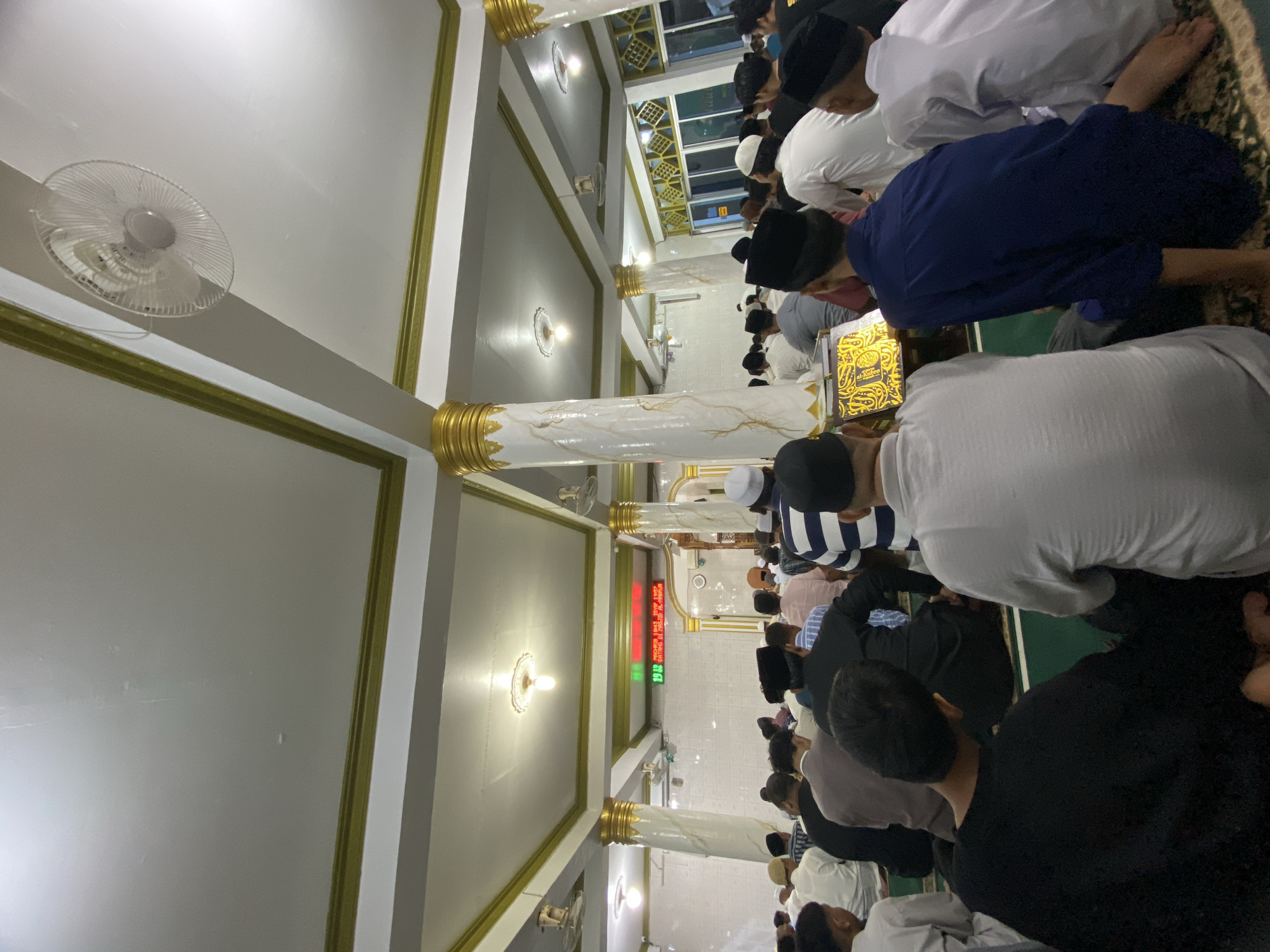 The image shows a large group of men praying inside a mosque. The mosque has white walls with gold trim and ornate columns. A digital sign in the background displays red and green numbers. The men are wearing a variety of clothing, including white robes and black caps. They are kneeling on a green carpet. The scene appears to be a regular prayer session in the city of Lhokseumawe, Indonesia.