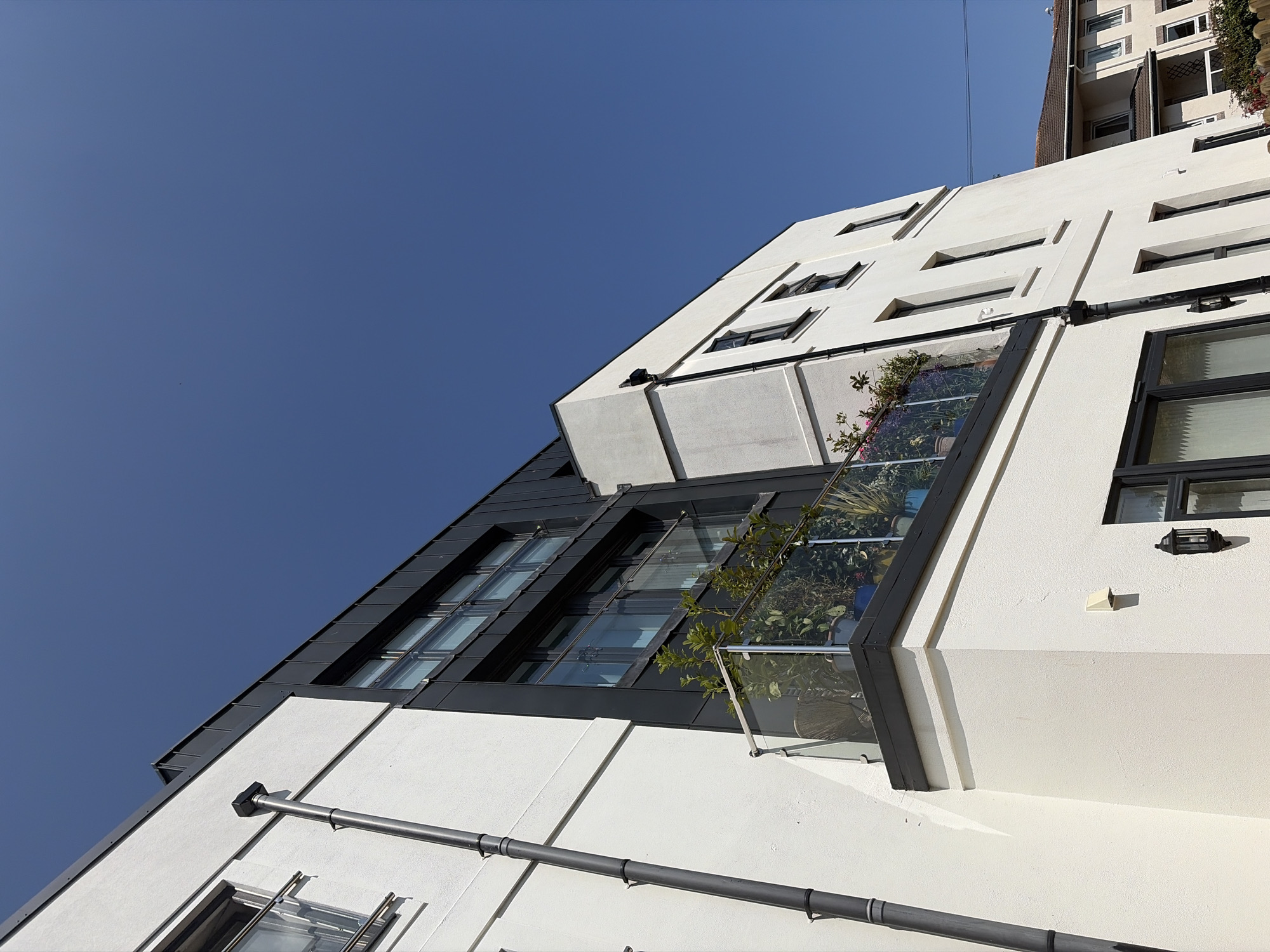 The image shows the exterior of a modern, white-rendered apartment building on a clear, sunny day. The building features dark-framed windows and a dark, angular roofline. There is a balcony with a glass balustrade, adorned with potted plants, extending from the upper floor. A dark drainpipe runs vertically down the side of the building. In the background, another residential building with a tiled roof is partially visible. The scene is set in Torquay, United Kingdom.